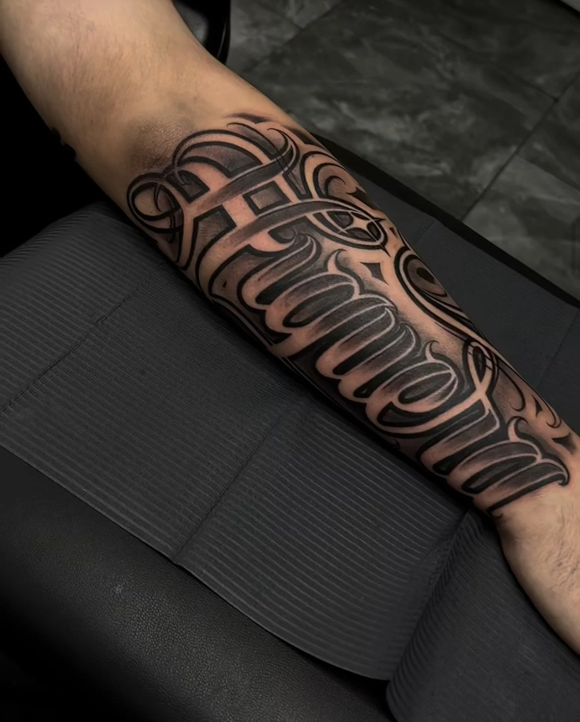 A person's arm with a large, intricate black tattoo with script lettering resting on a black textured surface.