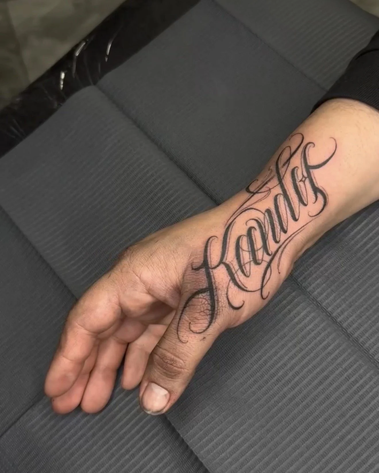 A person's arm with a tattoo of the word 'Faith' in cursive script on the forearm, shown resting on a gray textured surface.