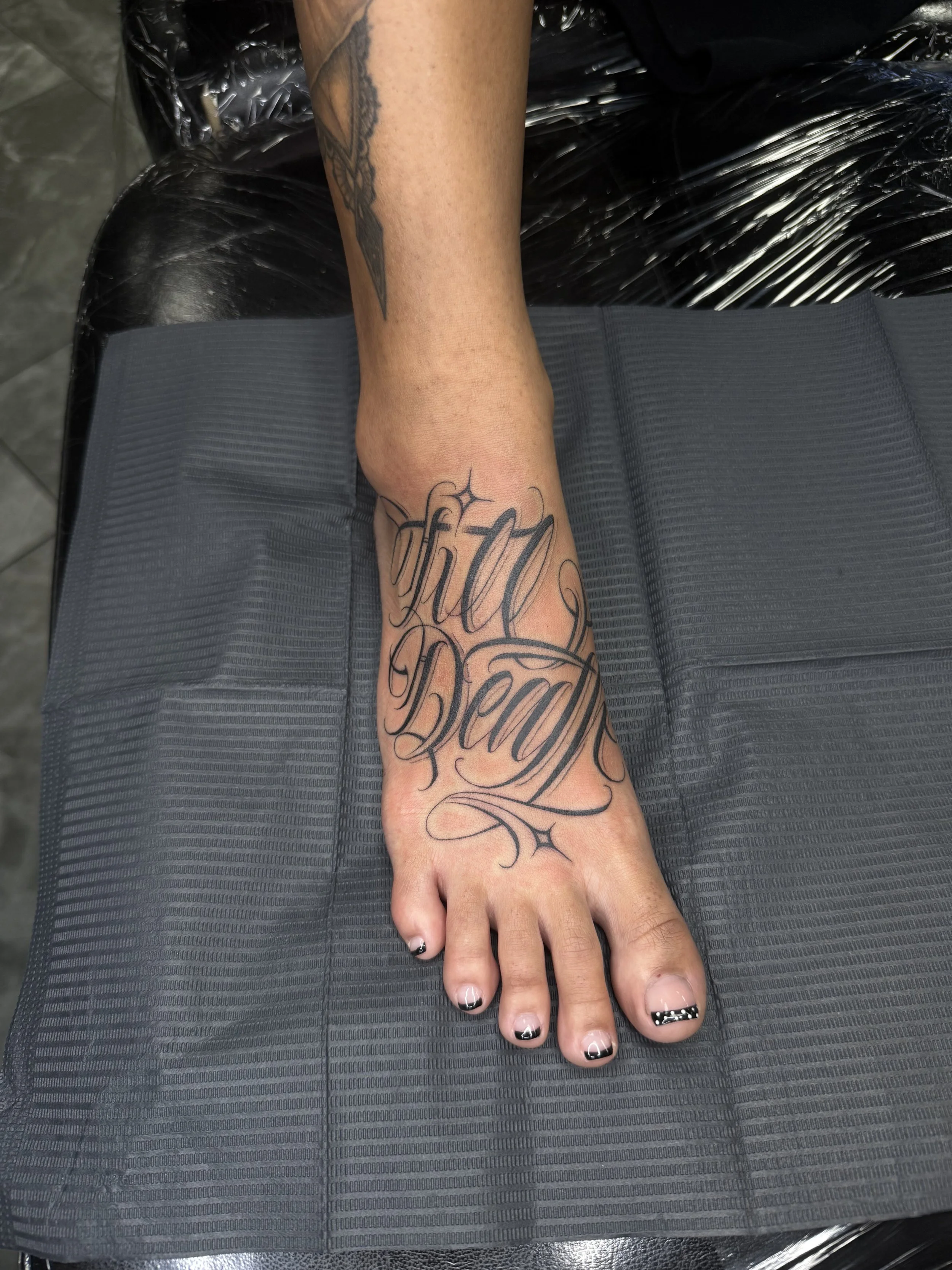 A person's foot with a tattoo that reads 'fifty' and 'death' in elaborate script, resting on a gray surface in a tattoo parlor.