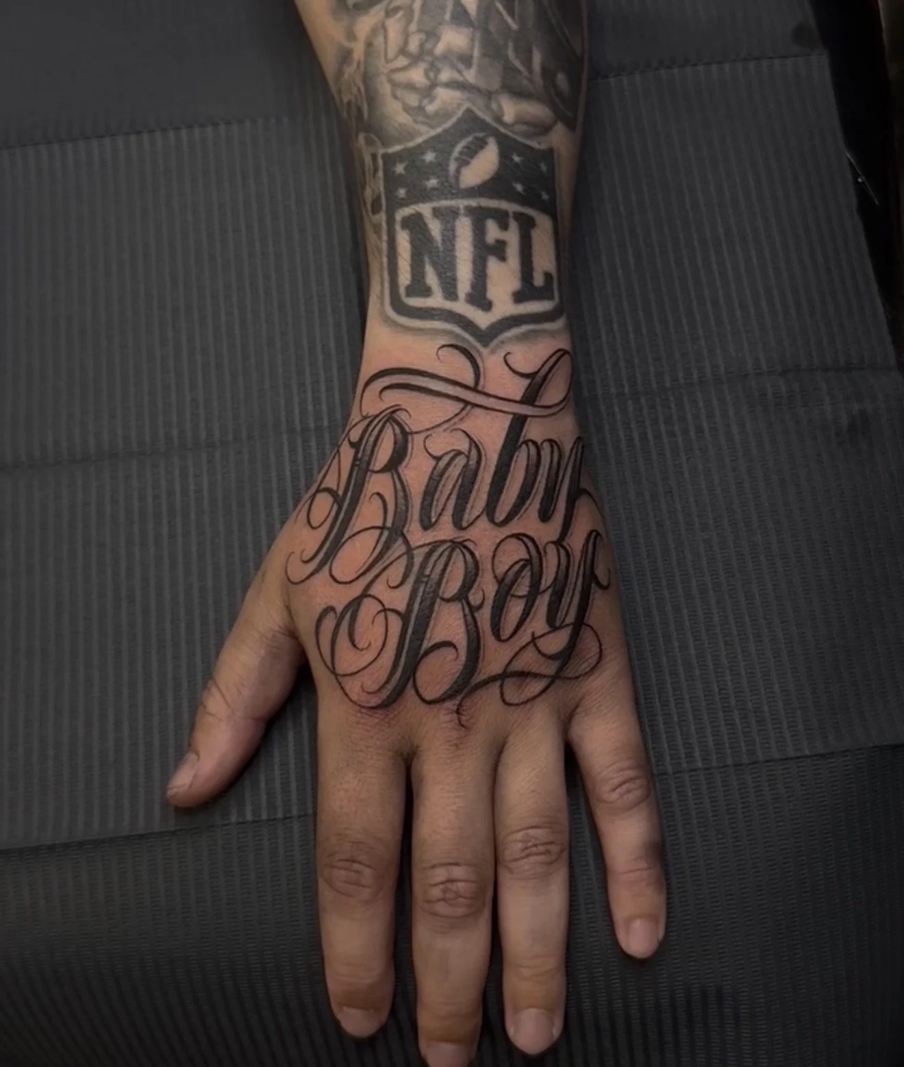 A person's hand with a tattoo that says 'Born Boy' in cursive script on the back of the hand. The wrist has an NFL logo tattoo above it, and part of an arm tattoo is visible above that.