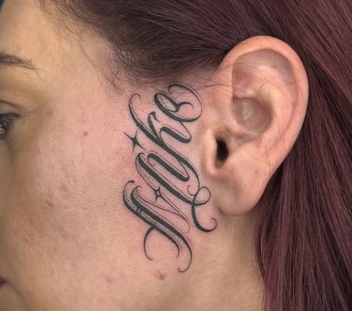 Tattoo of the word 'Blessed' in cursive script behind the right ear of a woman with reddish-brown hair.