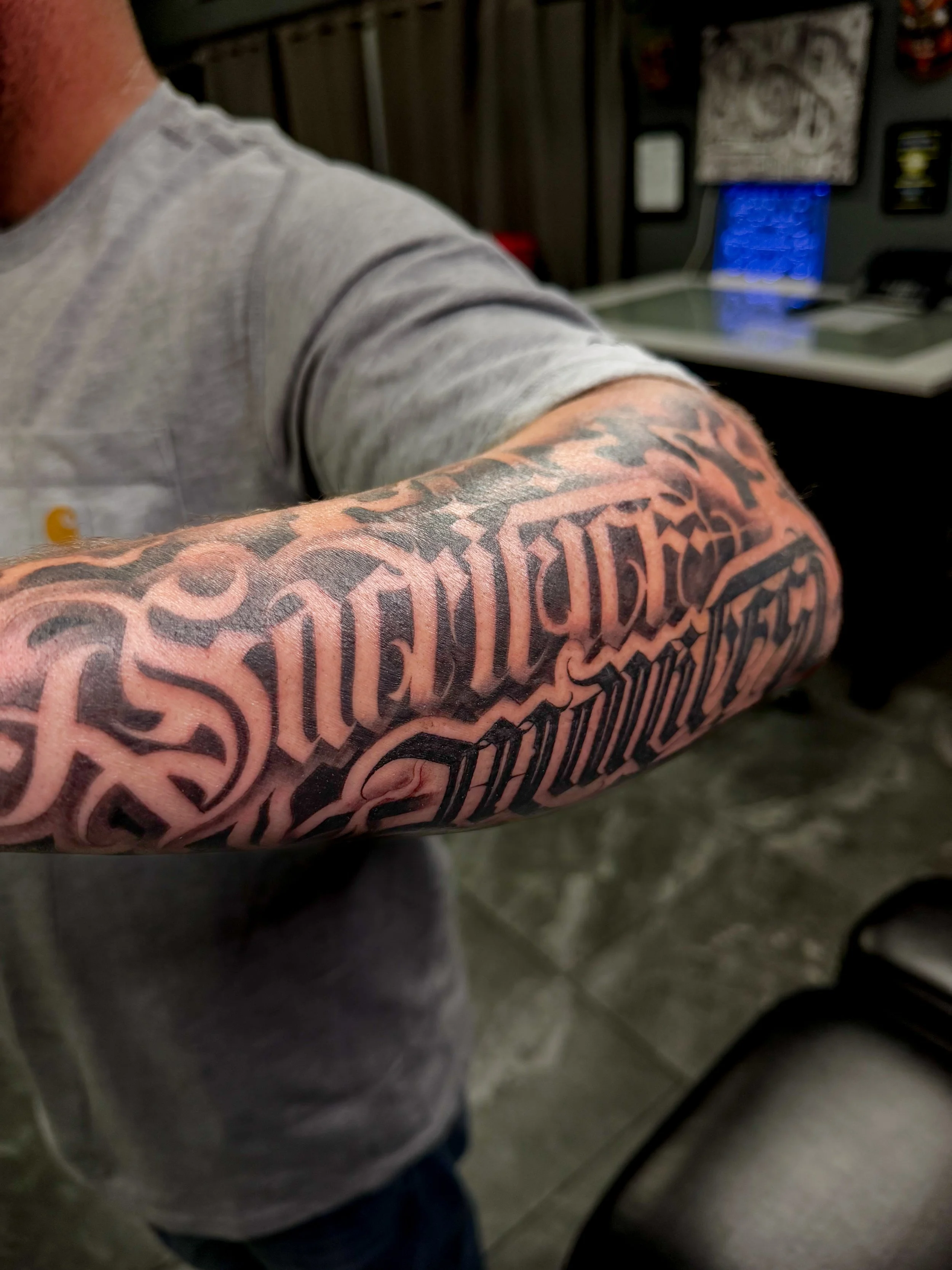 Close-up of a person's arm with a large black tattoo in gothic style lettering, wearing a gray short-sleeved shirt.