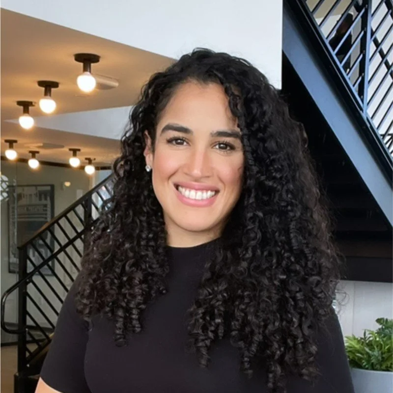 A woman with dark, curly hair smiling indoors near a modern staircase with black railing, white walls, and ceiling lights.