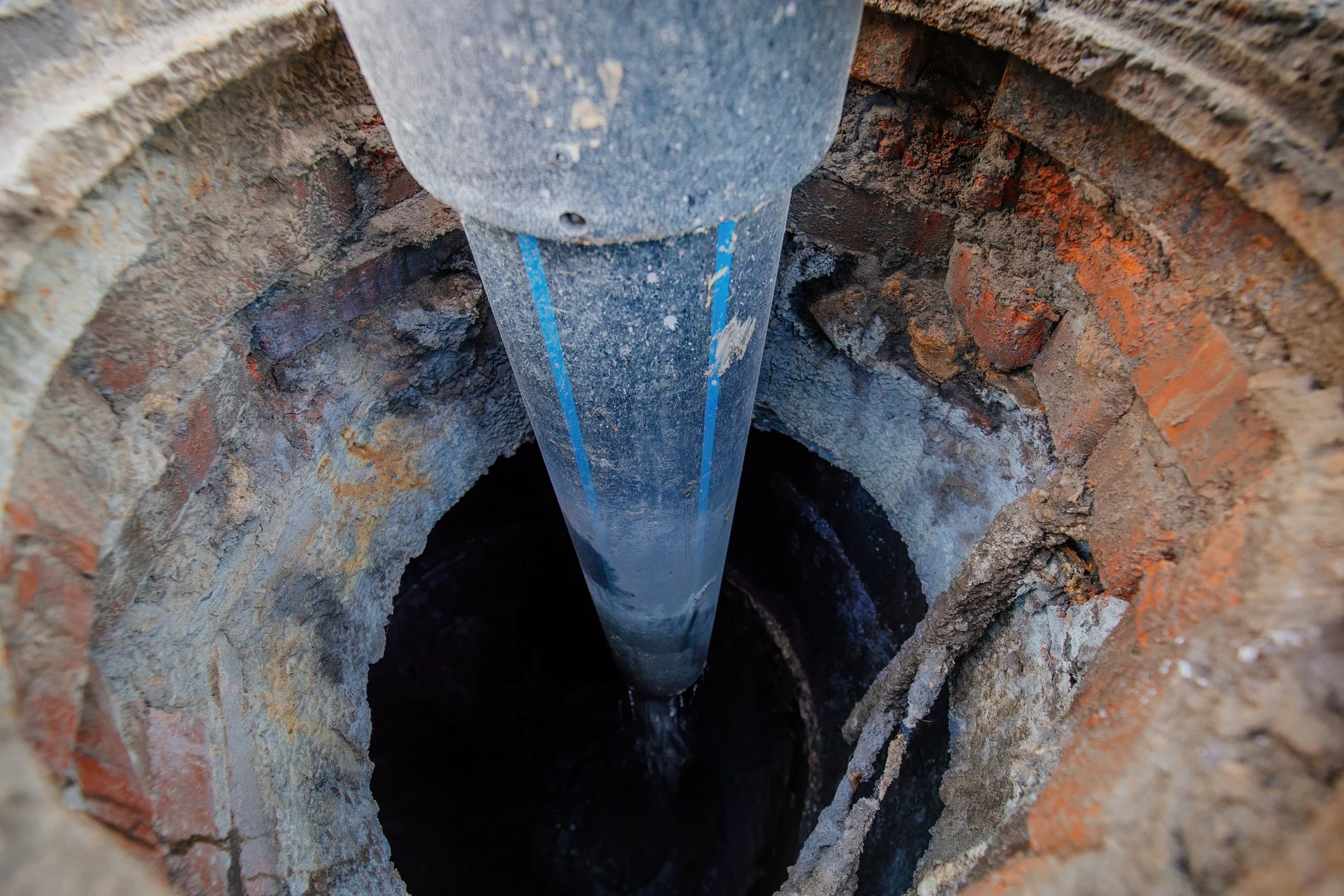 A close-up view of a manhole with a blue pipe leading down into it, surrounded by brick and concrete.