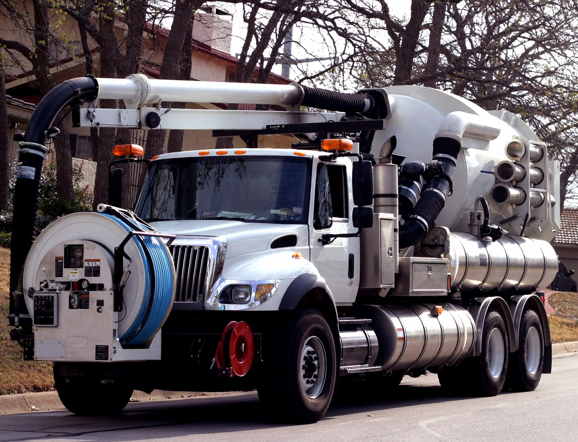A large vacuum excavator truck parked on a street with trees and houses in the background.