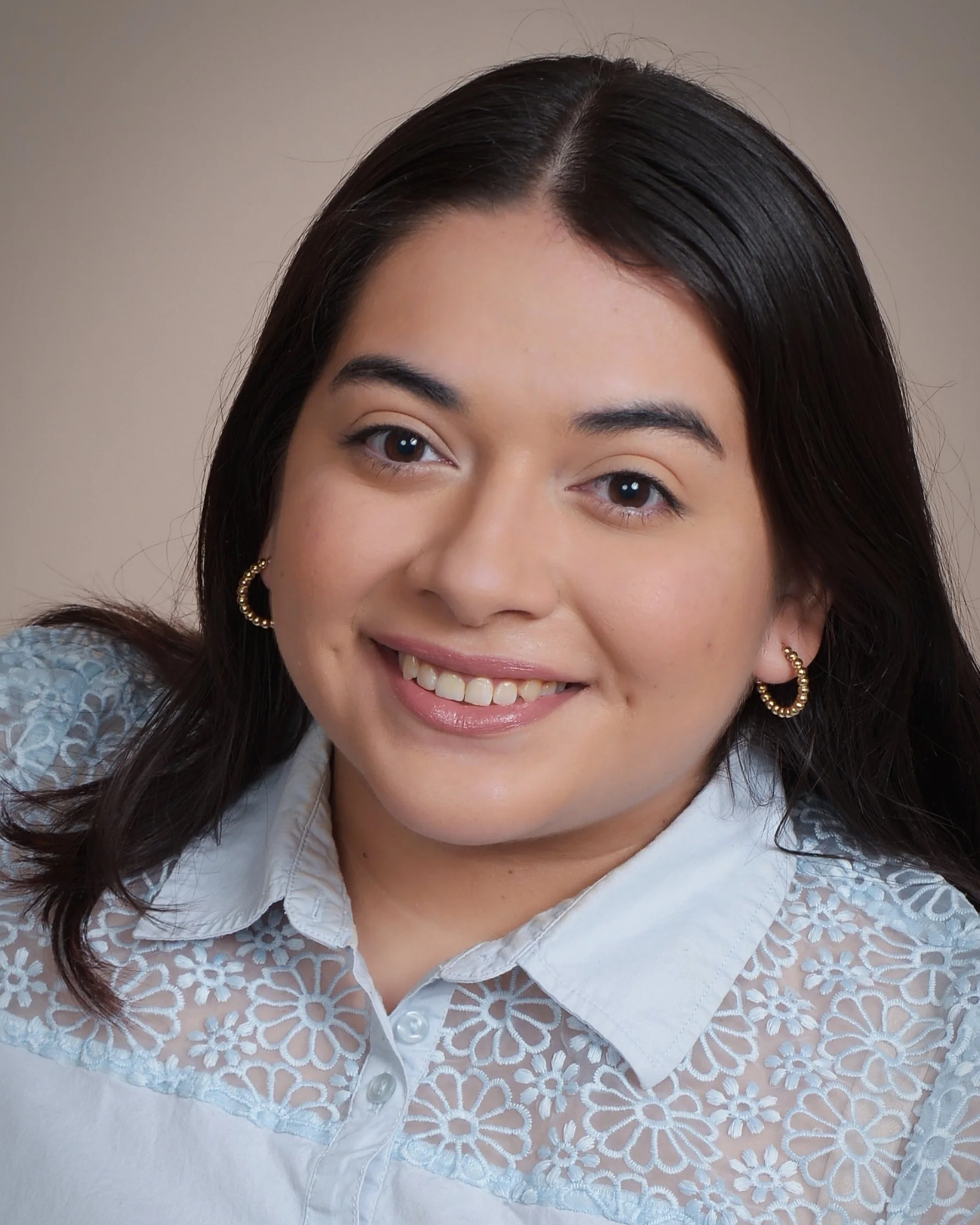 Close-up of a woman's face with a bright smile, dark hair, and gold hoop earrings, wearing a blue lace blouse.