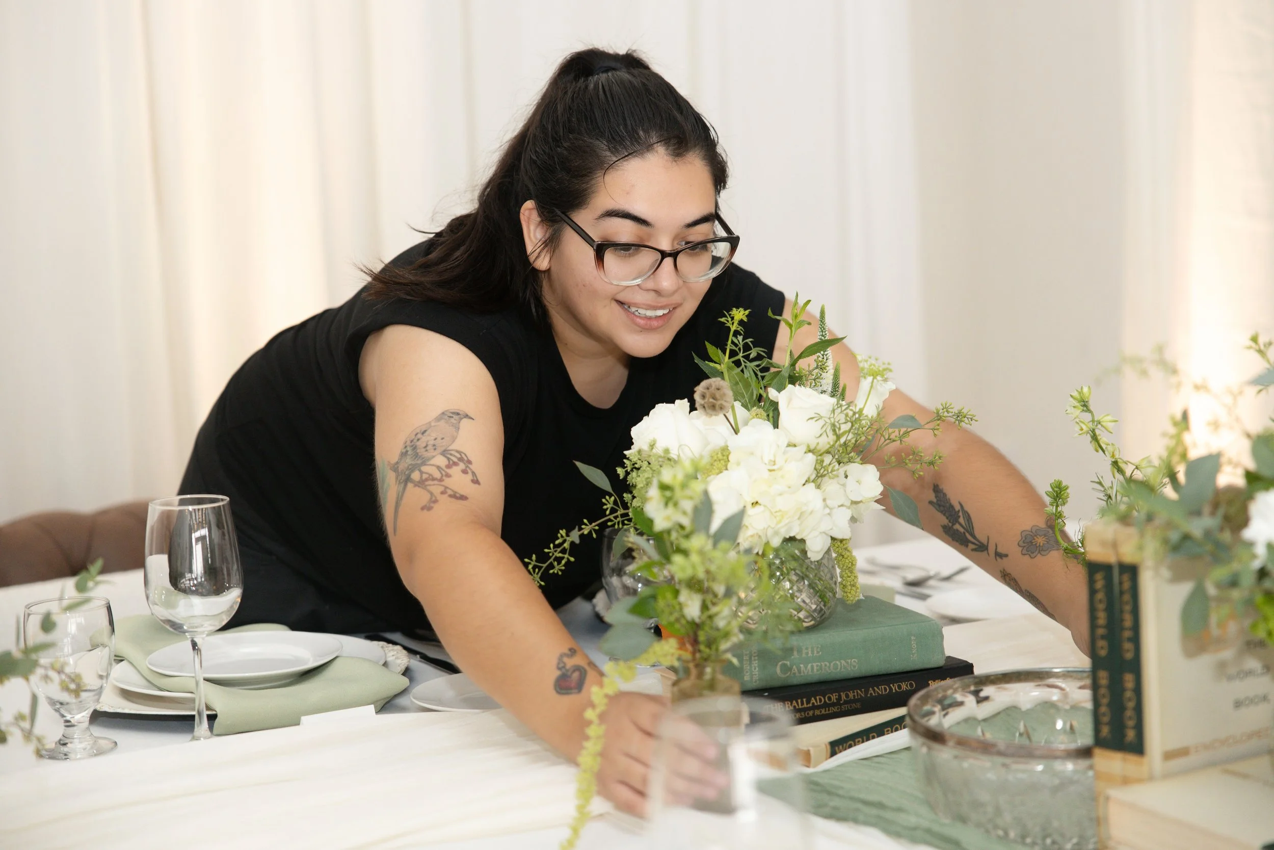 A wedding planner with tattoos is arranging a floral centerpiece at a table set for a meal, with books and candlelight around her.