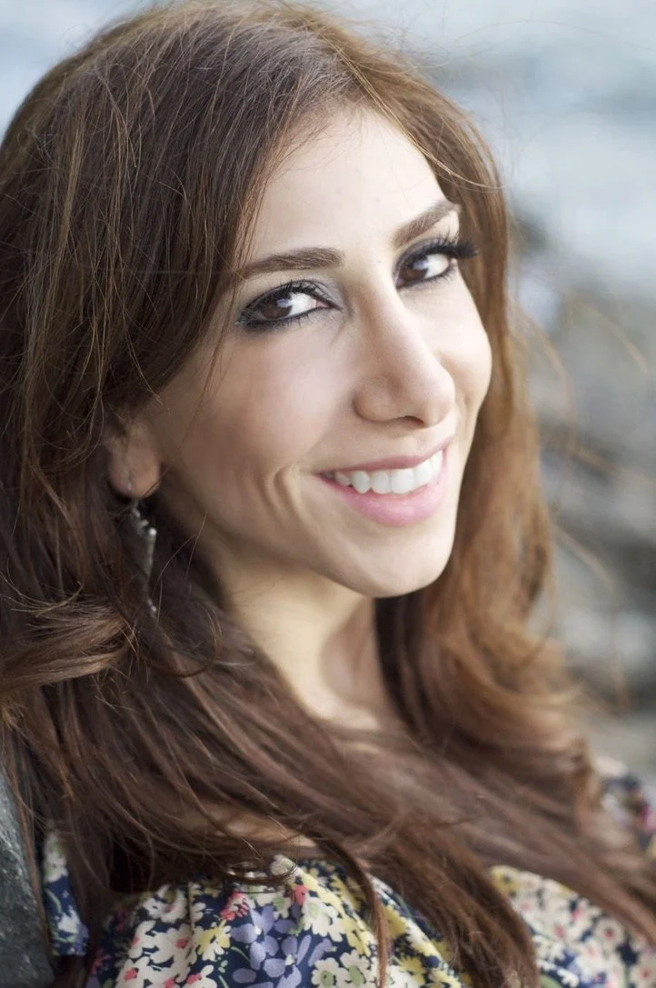 Close-up of a smiling woman with long, wavy, reddish-brown hair, wearing makeup and patterned clothing, outdoors with blurred background.