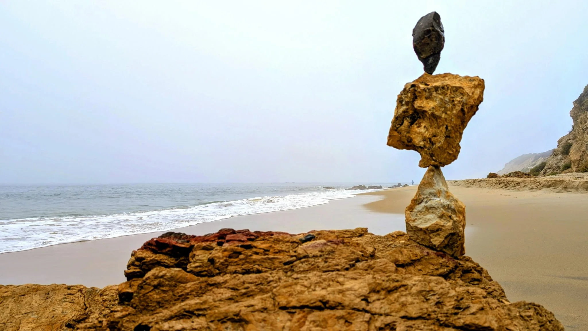 The image shows a beach with a stack of balanced rocks in the foreground, with the ocean and a hazy sky in the background.