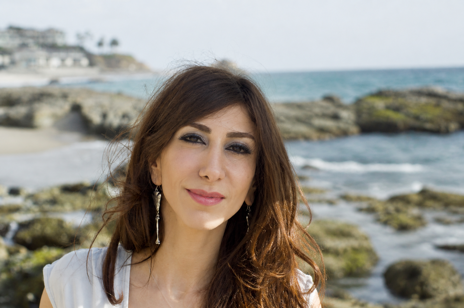 A woman with long brown hair and earrings smiling at the camera on a rocky beach with ocean waves and some houses on a hillside in the background.