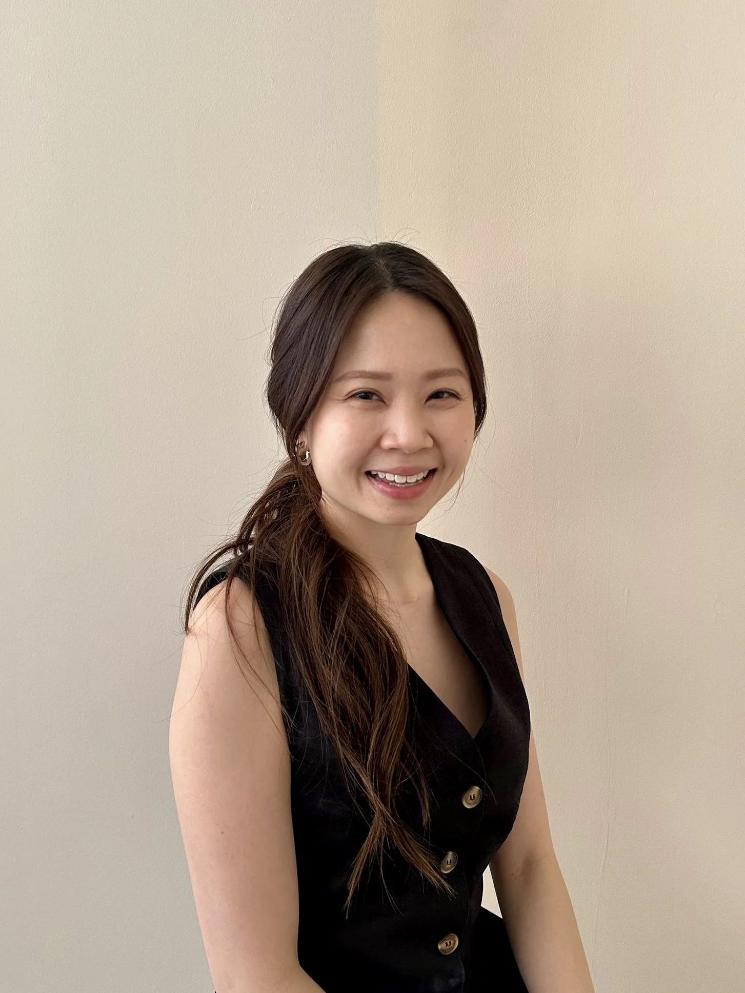 A young woman with long wavy brown hair smiling, wearing a black sleeveless dress with buttons on the front, standing against a plain light-colored wall.