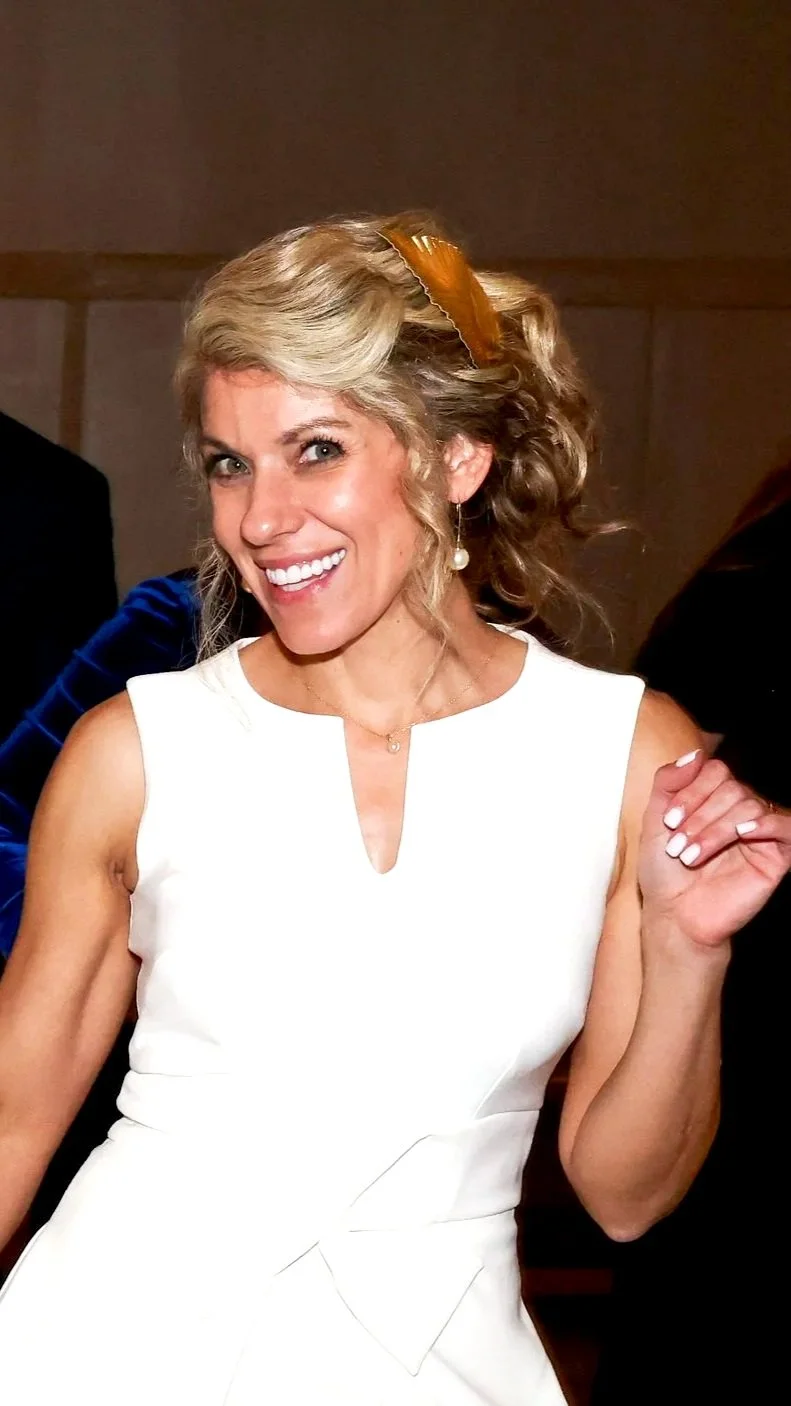 A woman with blonde, curly hair wearing a white sleeveless dress, smiling brightly, with pearl earrings and a peach-colored hair accessory.