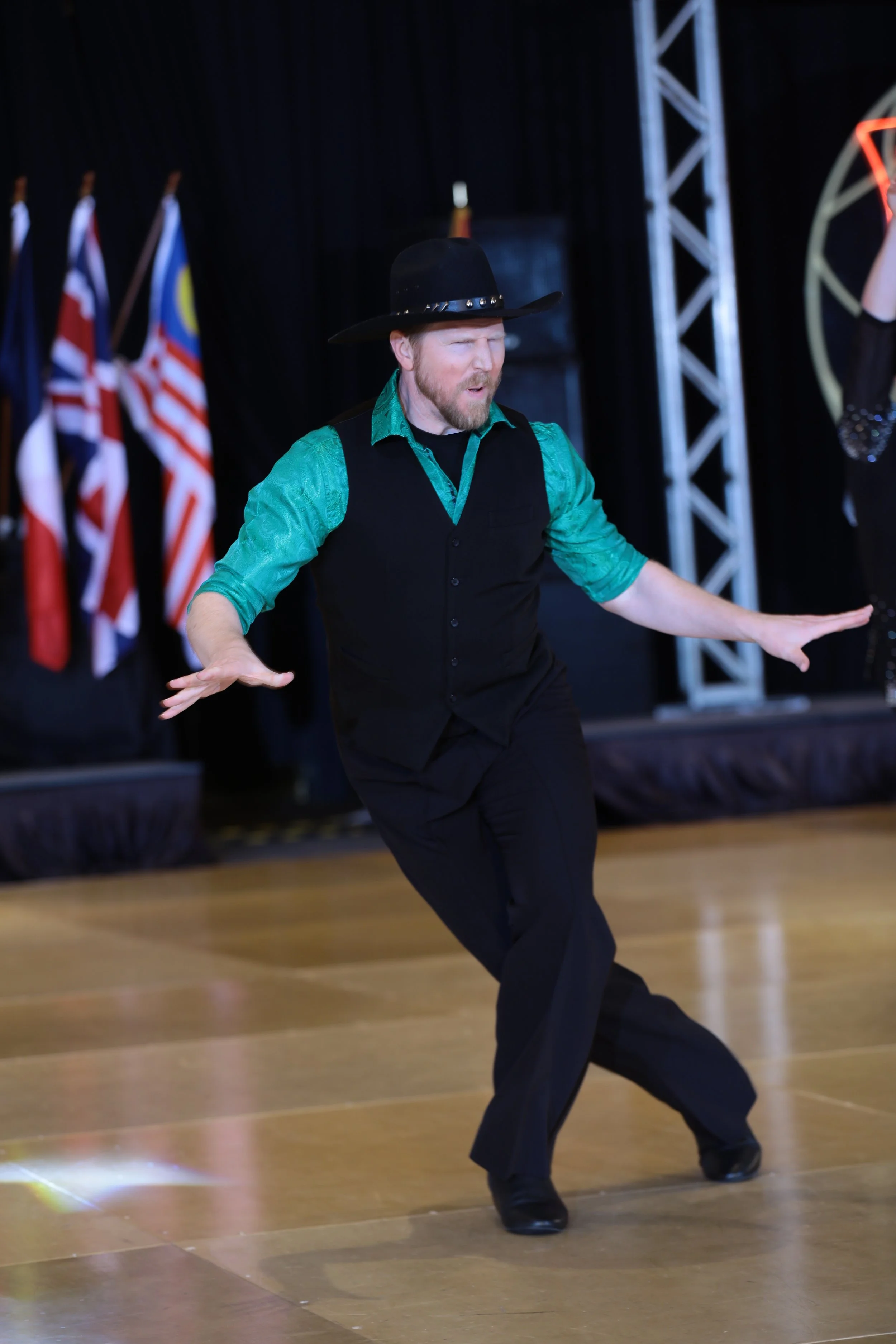 A man wearing a black cowboy hat, green shirt, black vest, and black pants is dancing on a wooden floor with a stage and flags in the background.