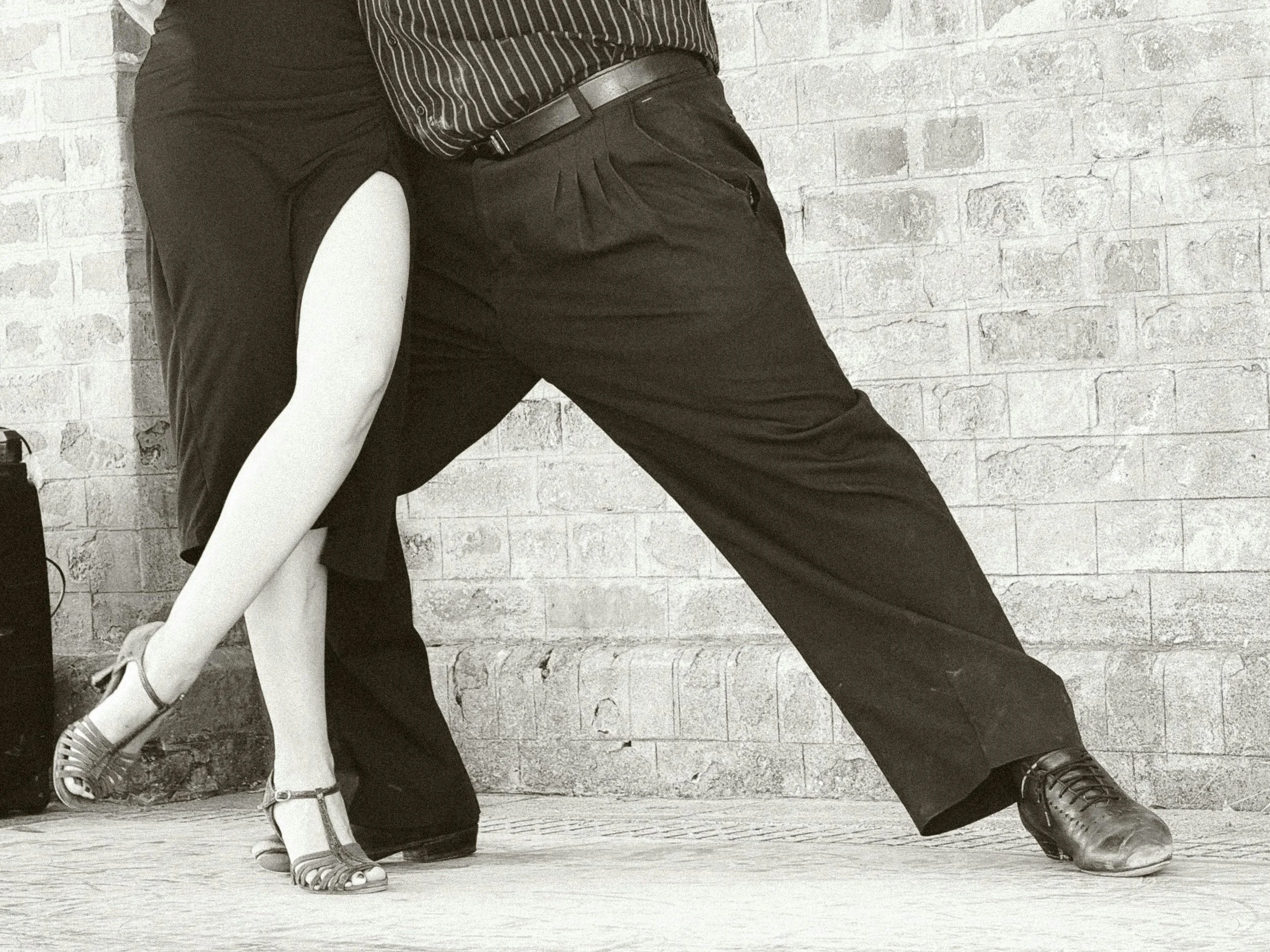 The image shows a close-up of two dancers' lower bodies, one wearing black pants and shoes, the other in a black and white striped shirt, black pants, and heels, performing in front of a brick wall.