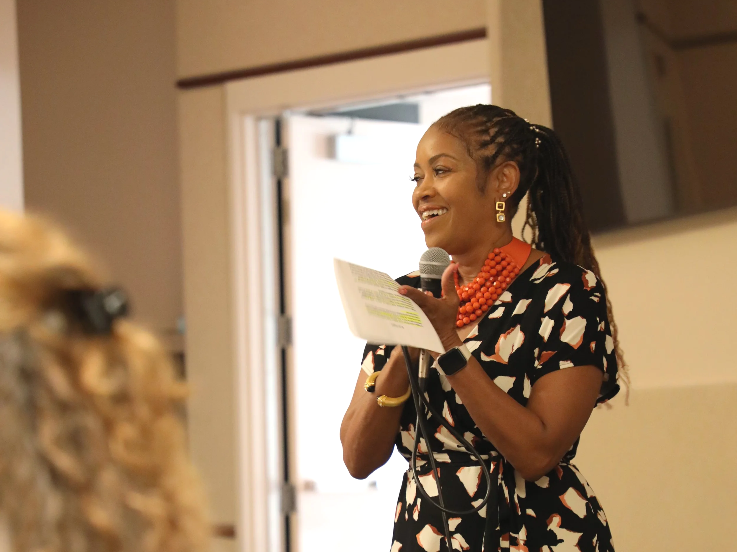 A woman with braided hair and earrings speaking into a microphone while holding a paper, standing in a room with a doorway and shelves in the background.
