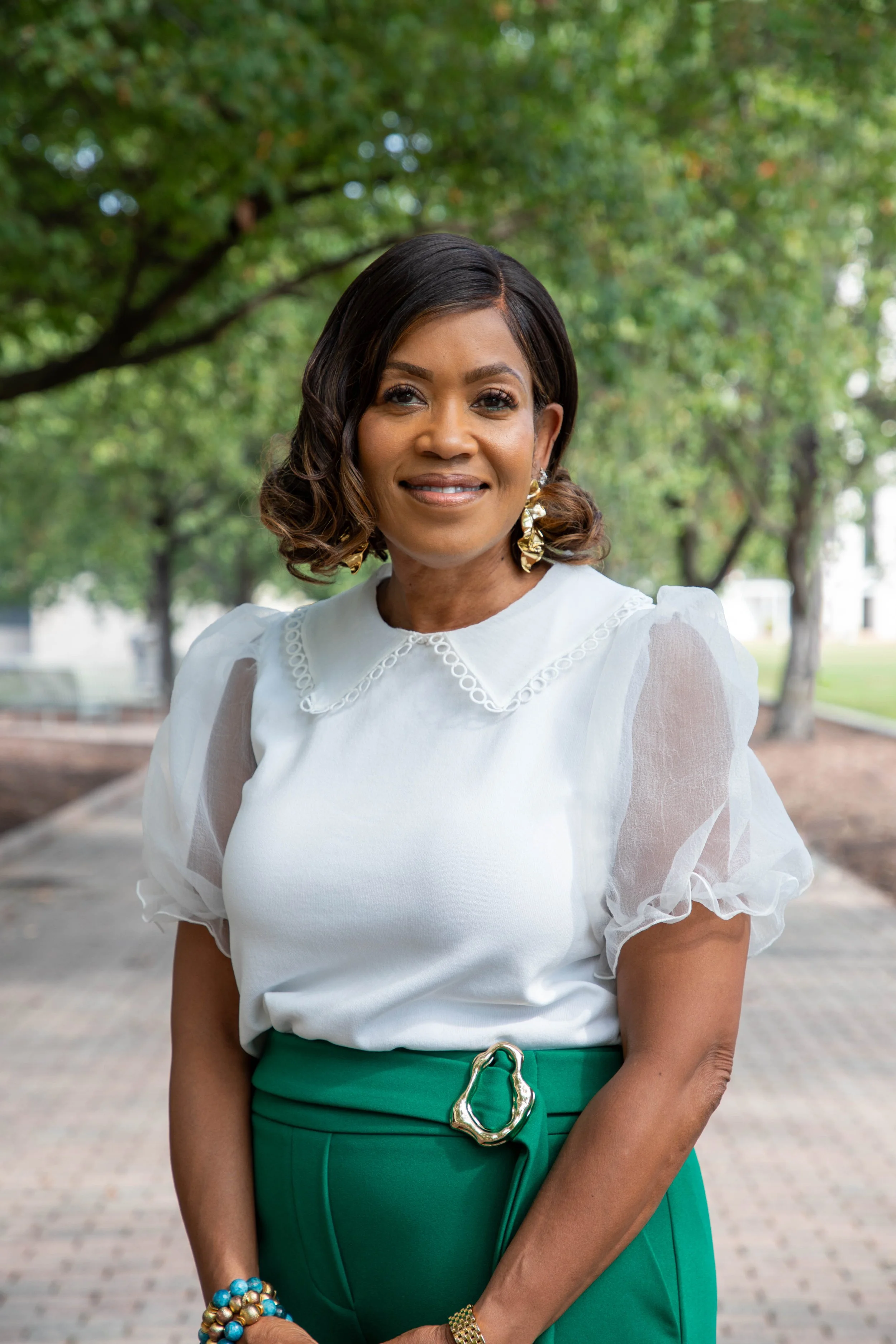 A woman with dark, shoulder-length hair smiling outdoors, wearing a white blouse with puffed sleeves, green high-waisted skirt with a decorative gold buckle, and colorful jewelry, in a park setting with trees in the background.
