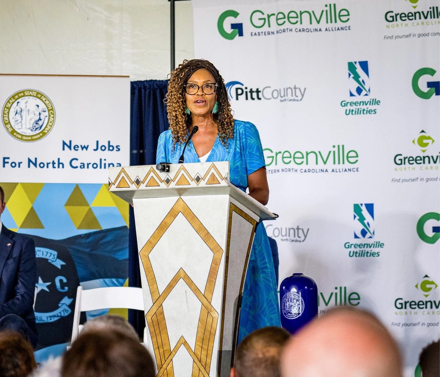 A woman in a blue dress with curly hair and glasses is speaking at a podium during a public event. Behind her are banners with logos and text promoting jobs in North Carolina and the Greenville area.