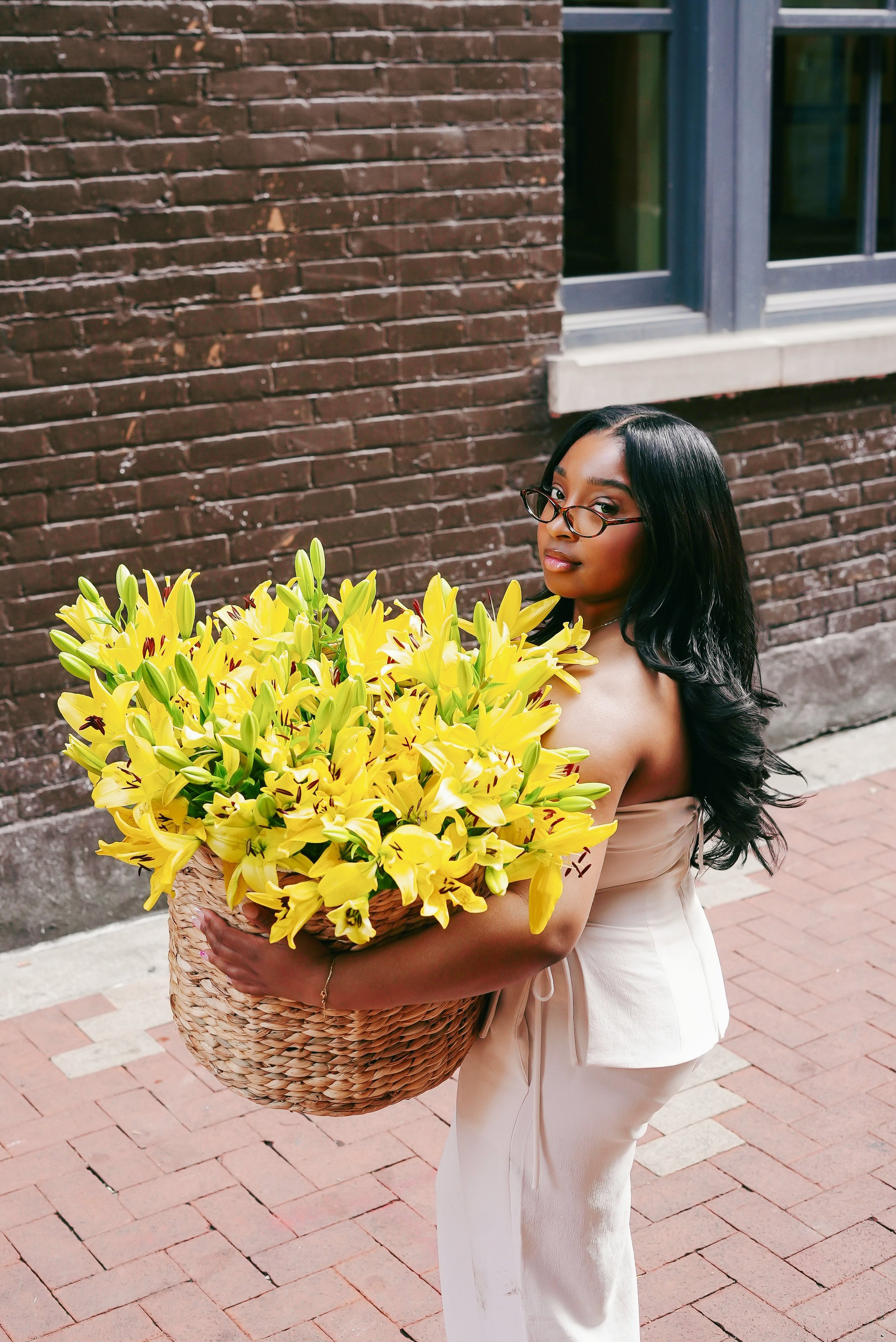 A woman with long black hair and glasses holding a large basket of yellow lilies on a brick sidewalk in front of a brick building.