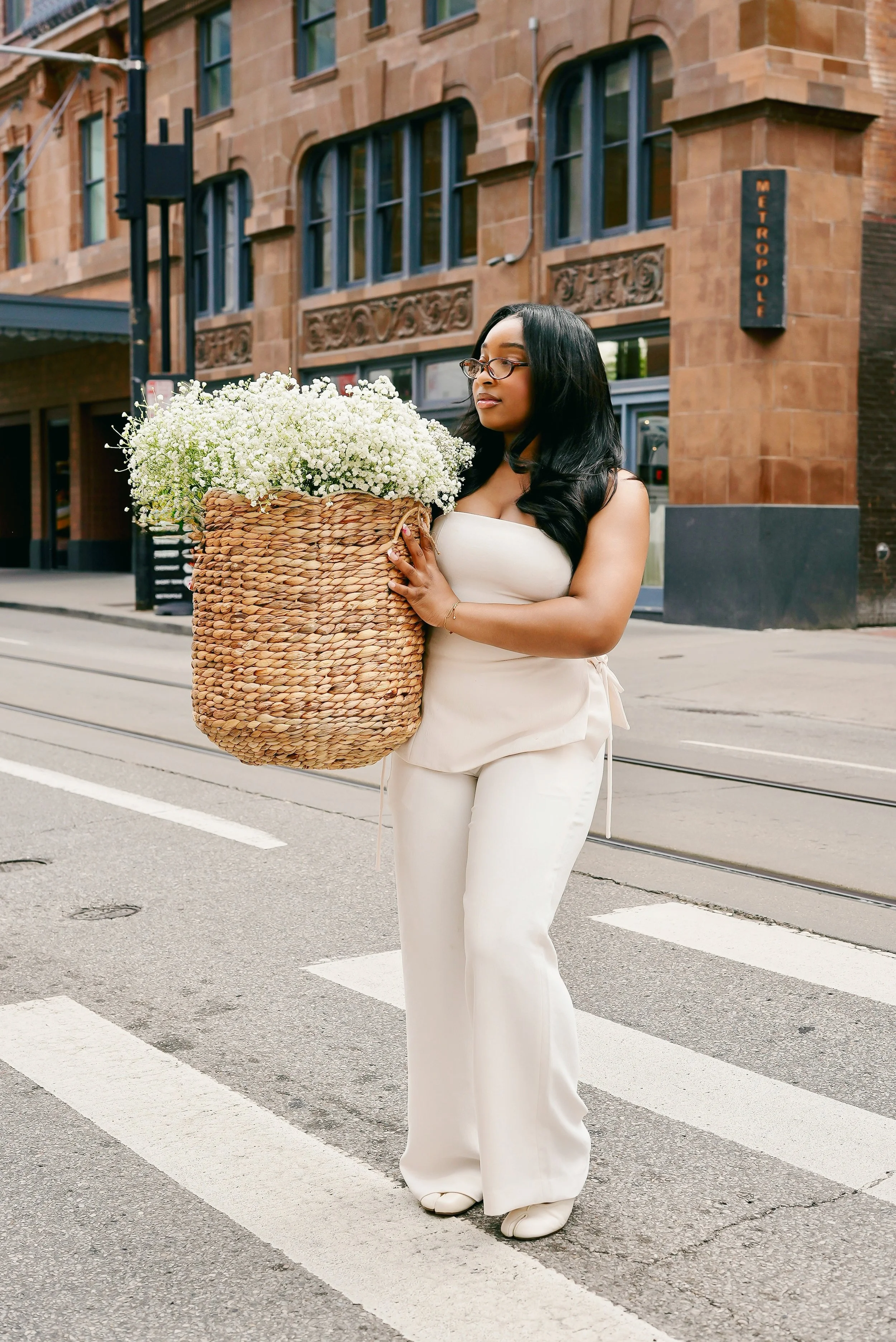 A woman with long black hair, glasses, and a cream-colored outfit holding a large woven basket filled with white flowers standing on a crosswalk in an urban setting.