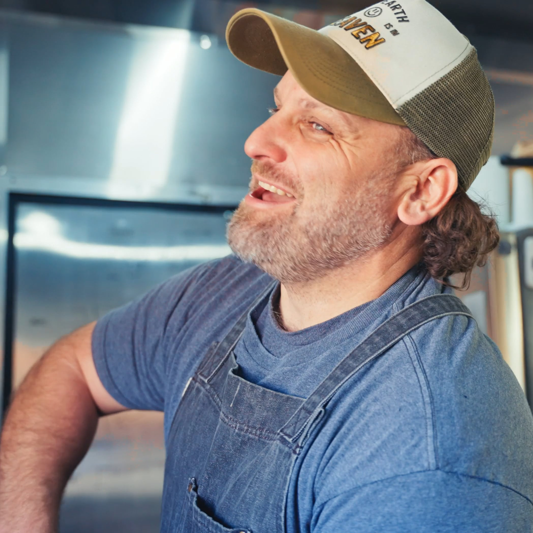 A man with a beard wearing a gray t-shirt, denim apron, and a beige and green trucker hat with logos and text, smiling in a kitchen setting.