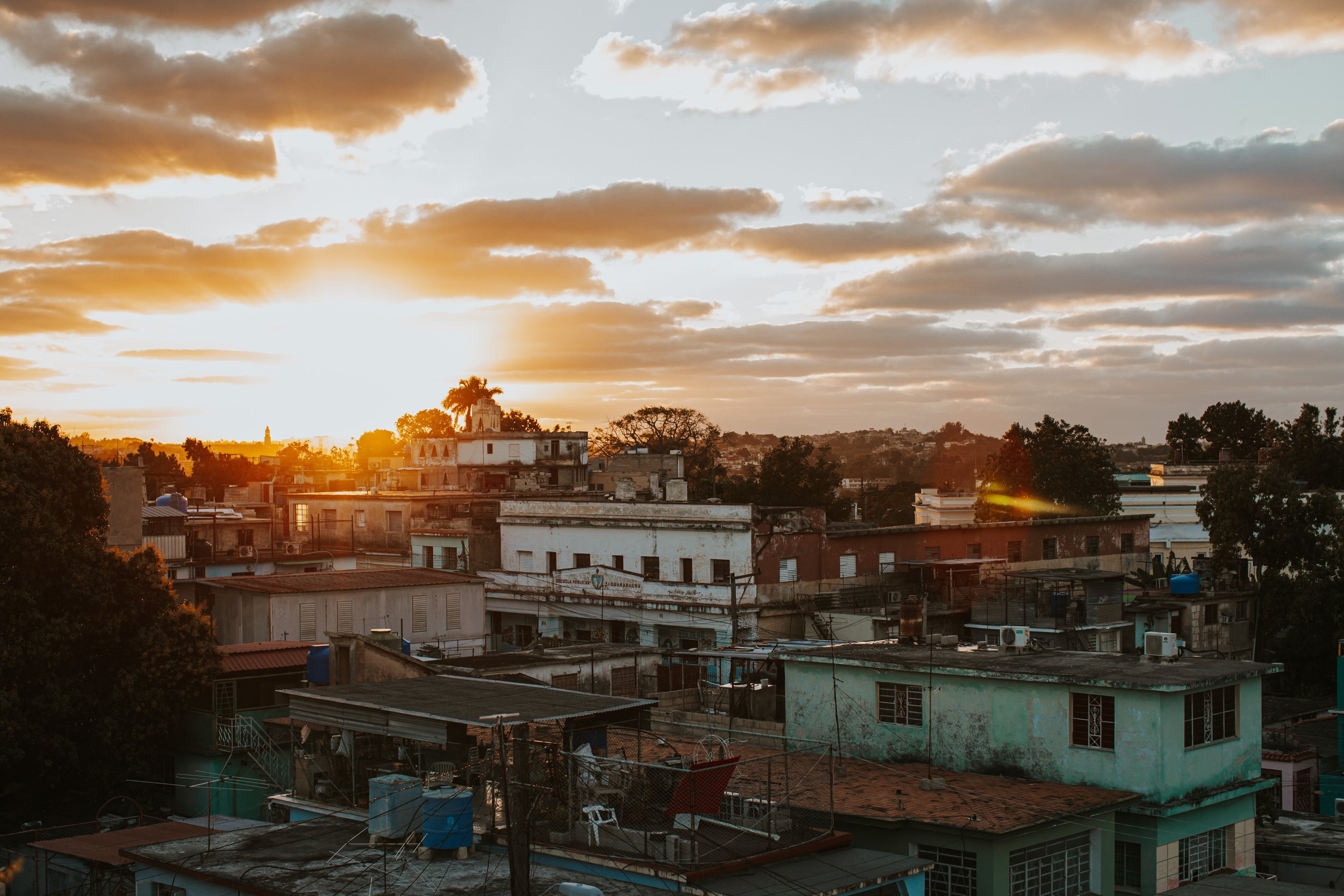 Sunset over a cityscape with low-rise buildings, trees, and clouds in the sky.