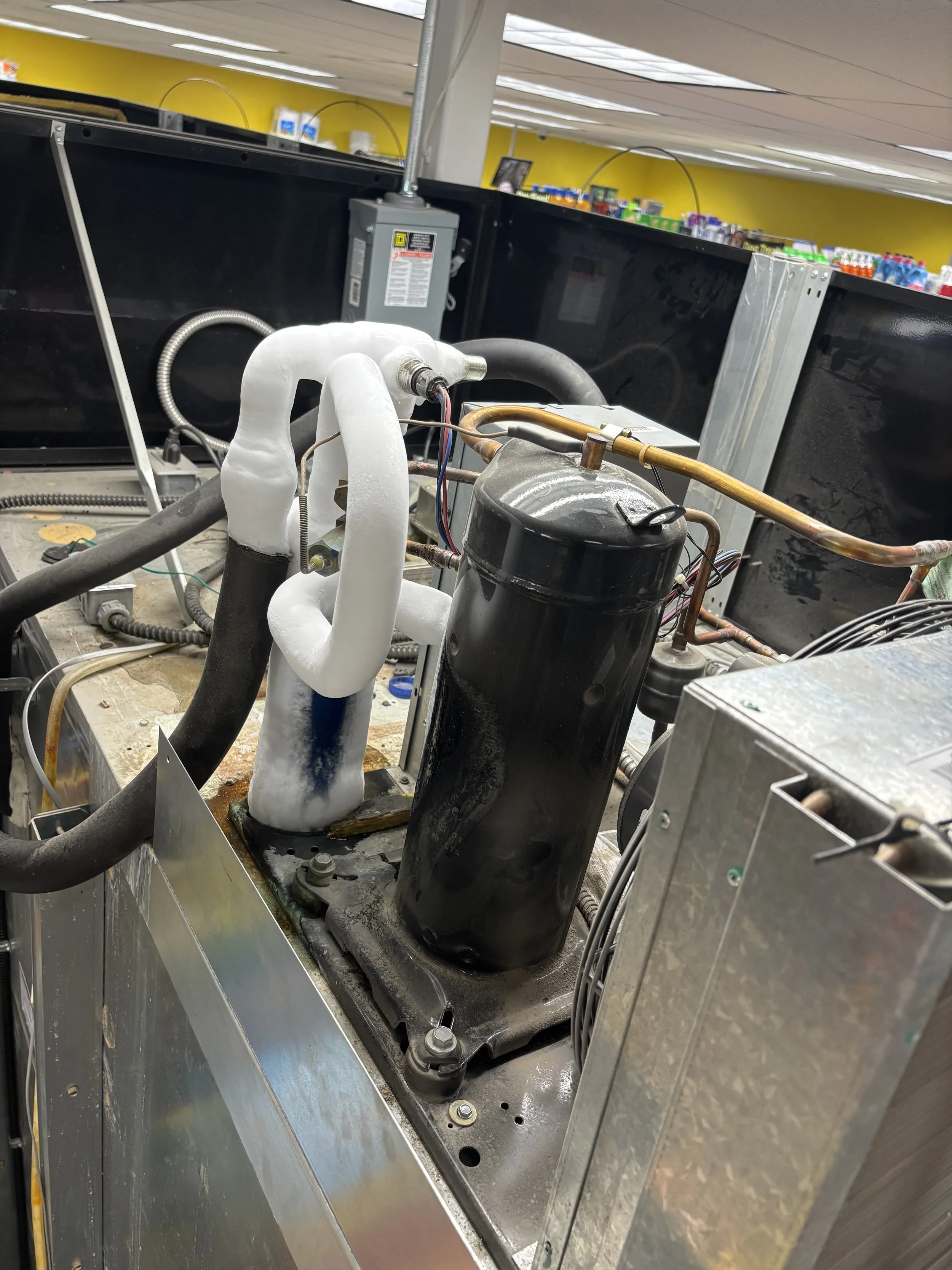 Close-up of an industrial HVAC or refrigeration system component, including a black compressor and copper piping, with some foam insulation around the pipes, in a commercial setting.