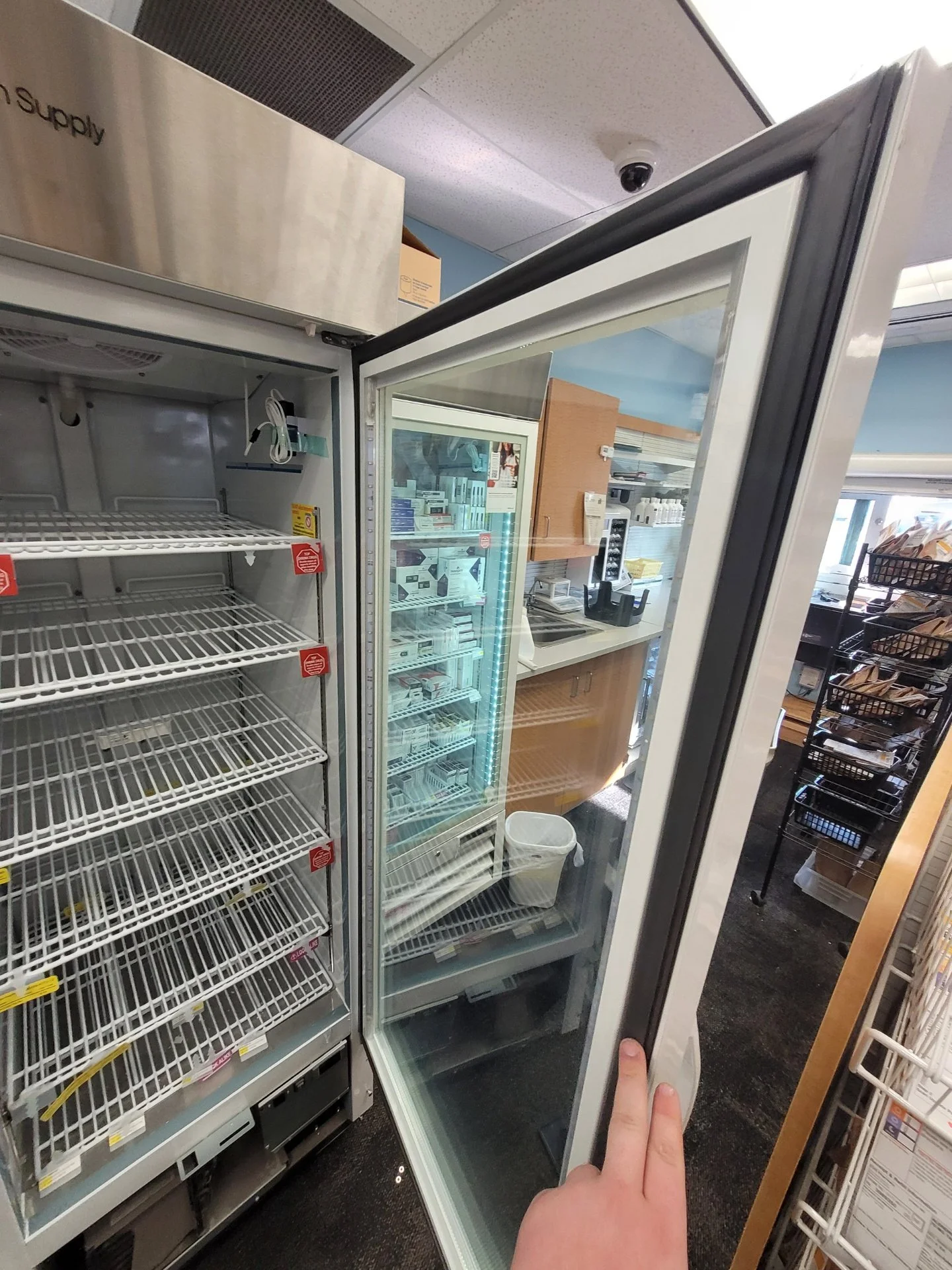 Open glass door of a commercial refrigerator in a store, with empty wire shelves inside and a hand holding the door open.