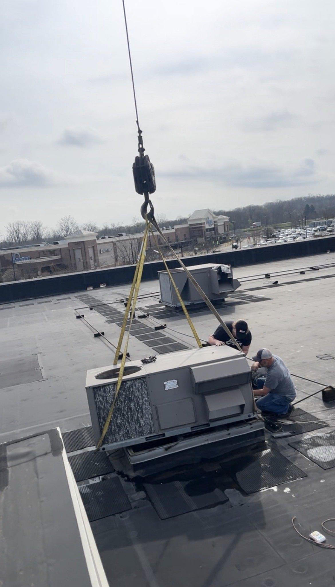 Two workers installing or repairing an HVAC unit on a commercial building's roof, with a crane and safety straps supporting the unit.