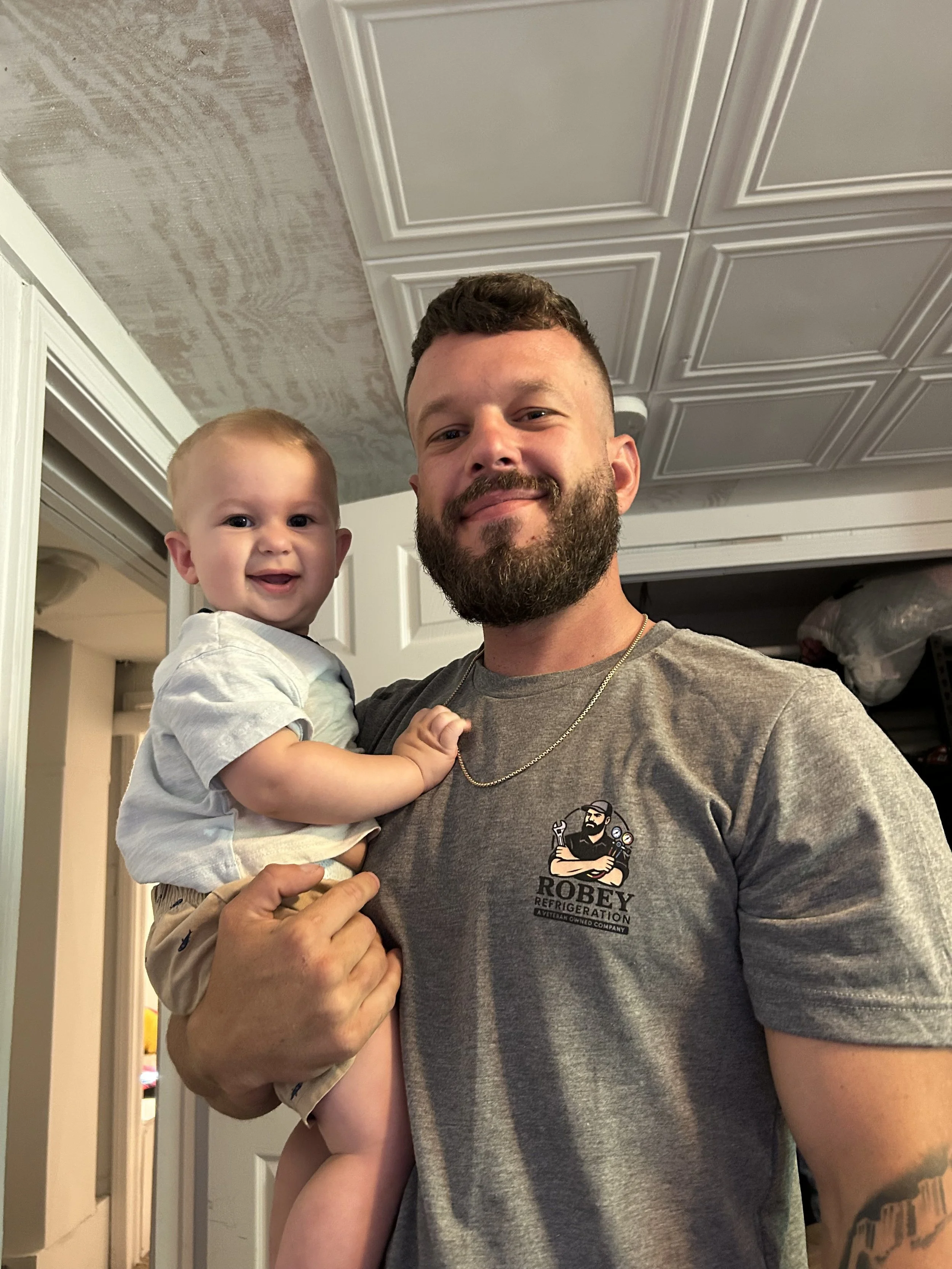 A man with a beard holding a smiling toddler with blonde hair in a house, both looking at the camera.