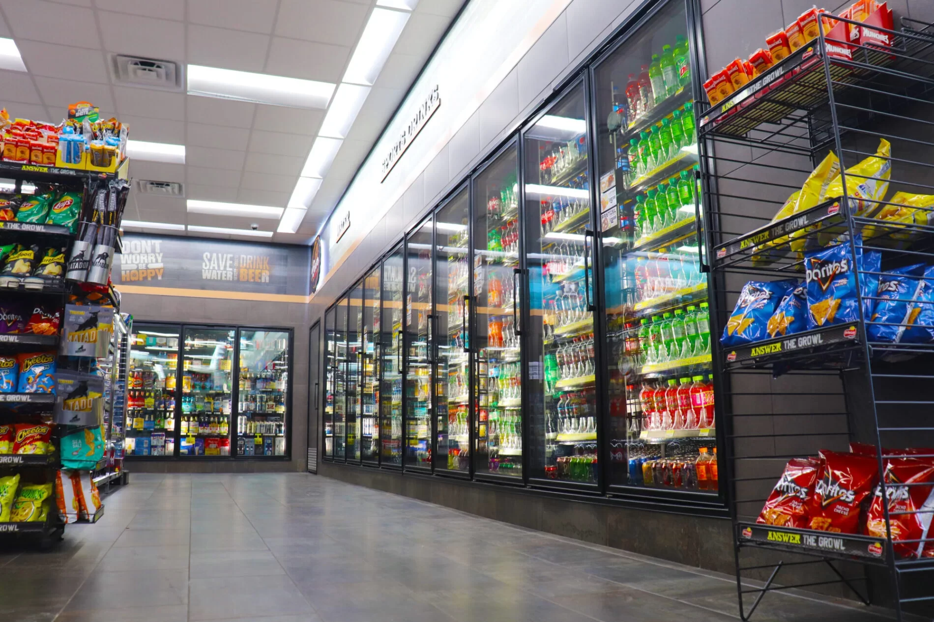 Inside a convenience store aisle with glass-door walk in refrigerator stocked with soft drinks and shelves with snack chips