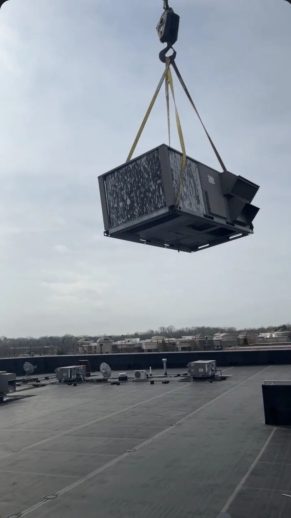 A large HVAC unit is being hoisted by a crane on the rooftop of a building, with clouds in the sky and other rooftop equipment visible below.