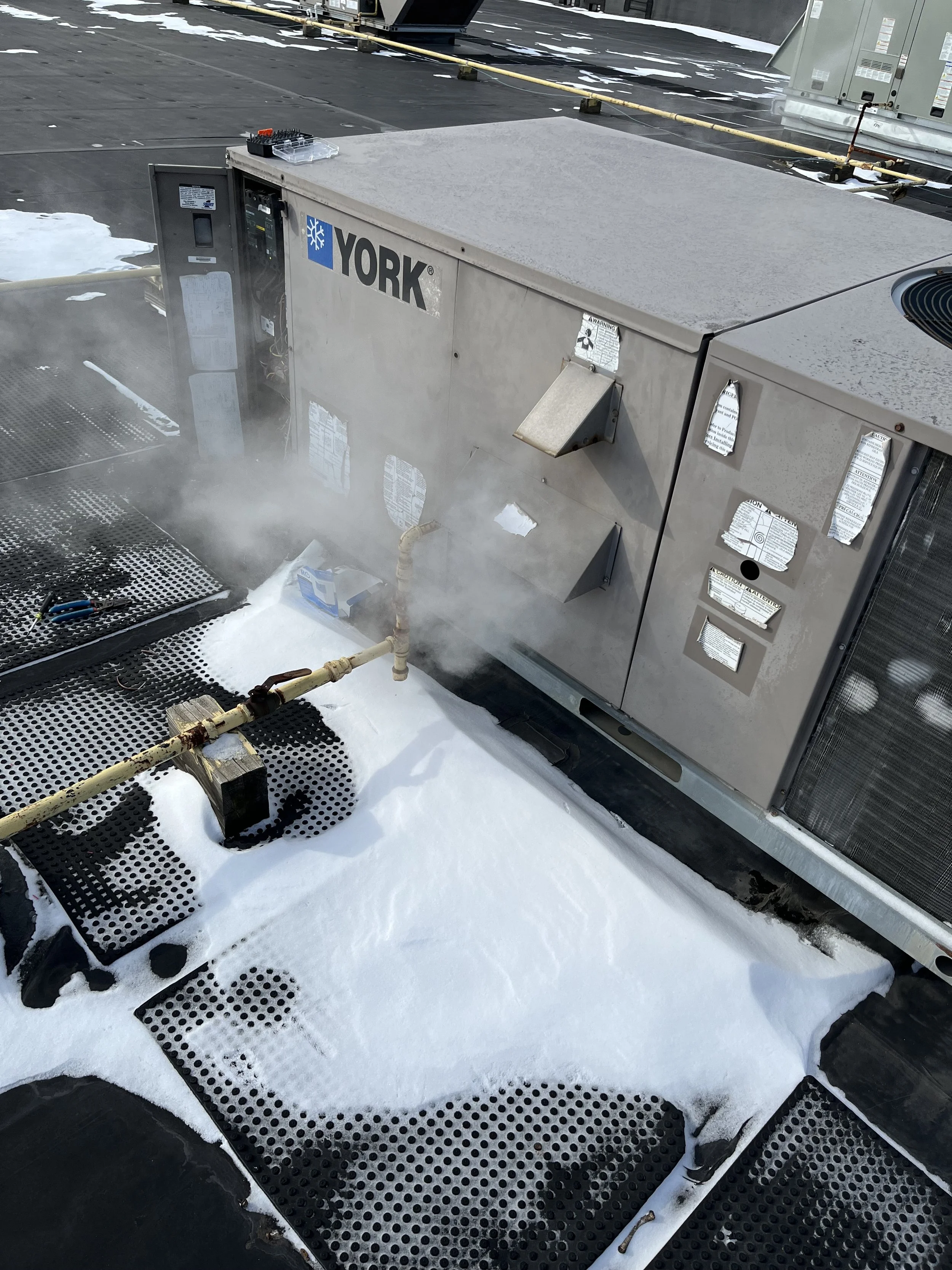 Air conditioning unit on a rooftop emitting steam in snowy weather, with a partially melted snow patch and black rubber mats on the surface.