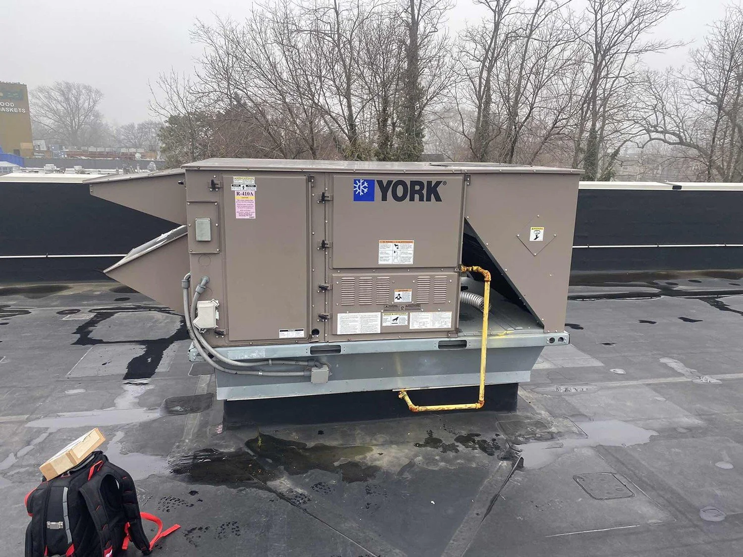 HVAC rooftop air conditioning unit labeled York installed on flat rooftop with a backpack and a package on the ground nearby, overcast sky with bare trees in the background.