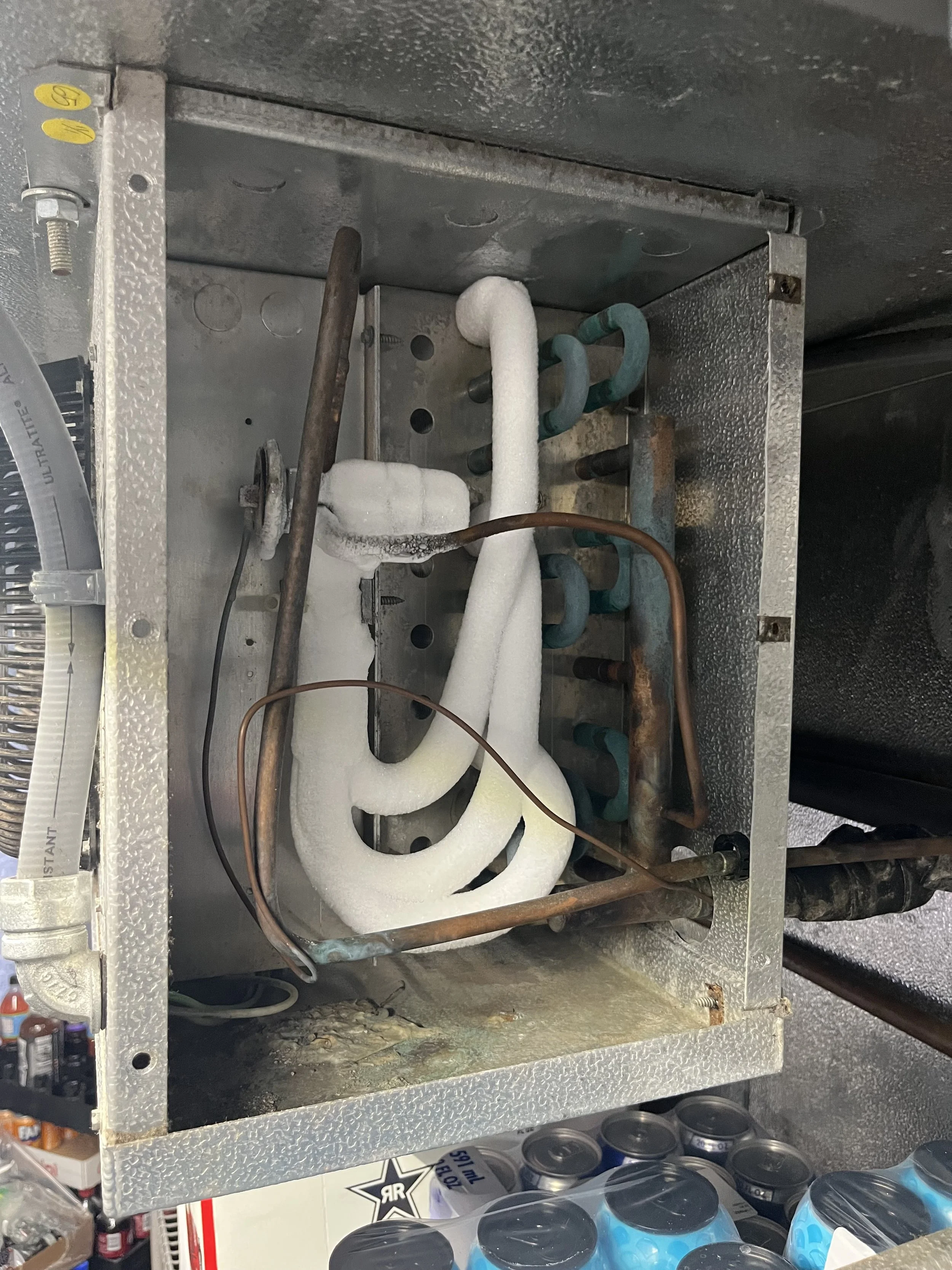 Interior of an old refrigeration or air conditioning unit with exposed metal components, copper and aluminum pipes, some rusted, with insulation and wiring, and cans of water below.
