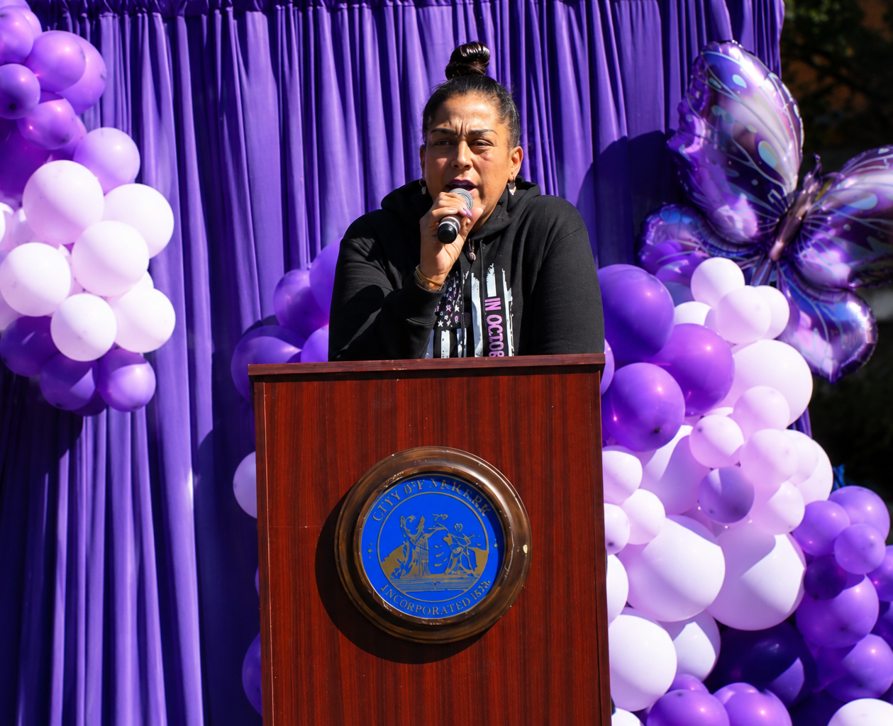 A woman speaking at a podium during a public event or rally, with purple balloons and a purple curtain backdrop behind her.