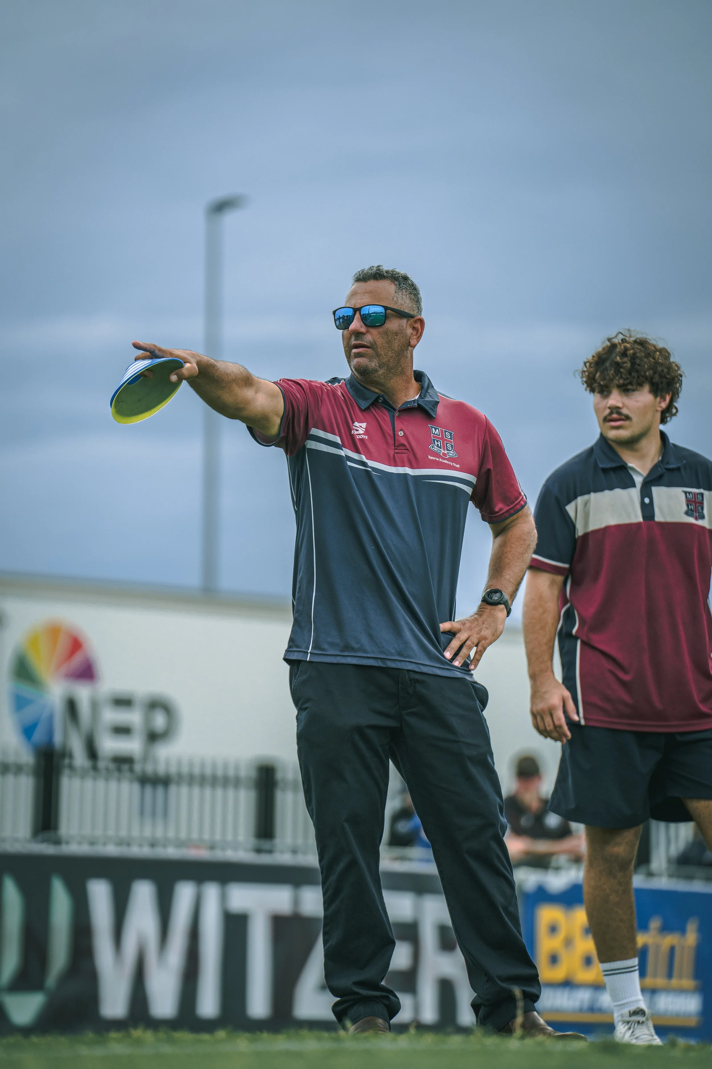 A man wearing sunglasses and a sports shirt is pointing with his right hand while two young men stand nearby. They are on a sports field, possibly during a game or practice, with a cloudy sky overhead.