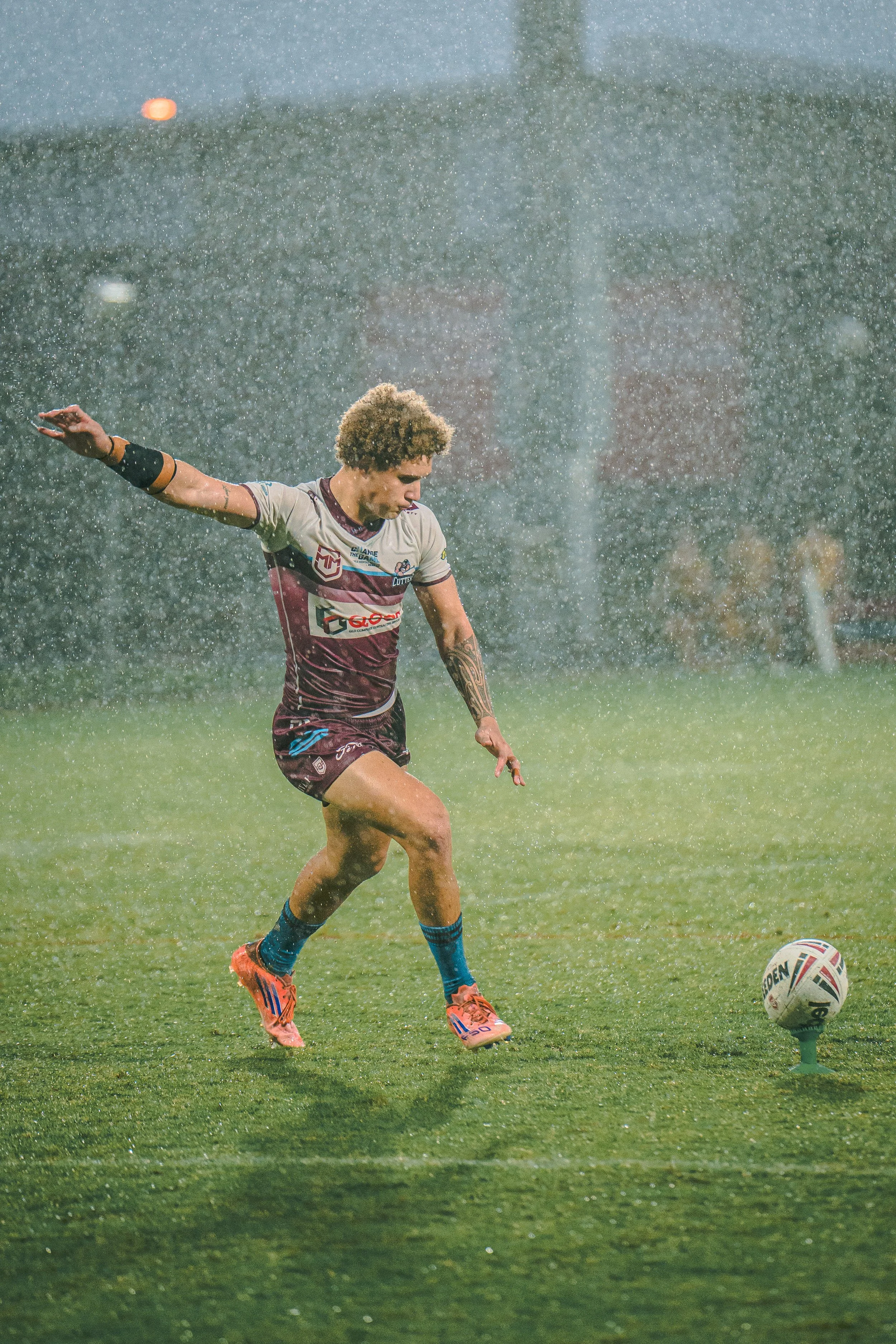 A rugby player in a grey and maroon uniform with tattoos on his right arm, wearing orange cleats and rolling blue socks, kicks a white rugby ball during a rainstorm on a grassy field.