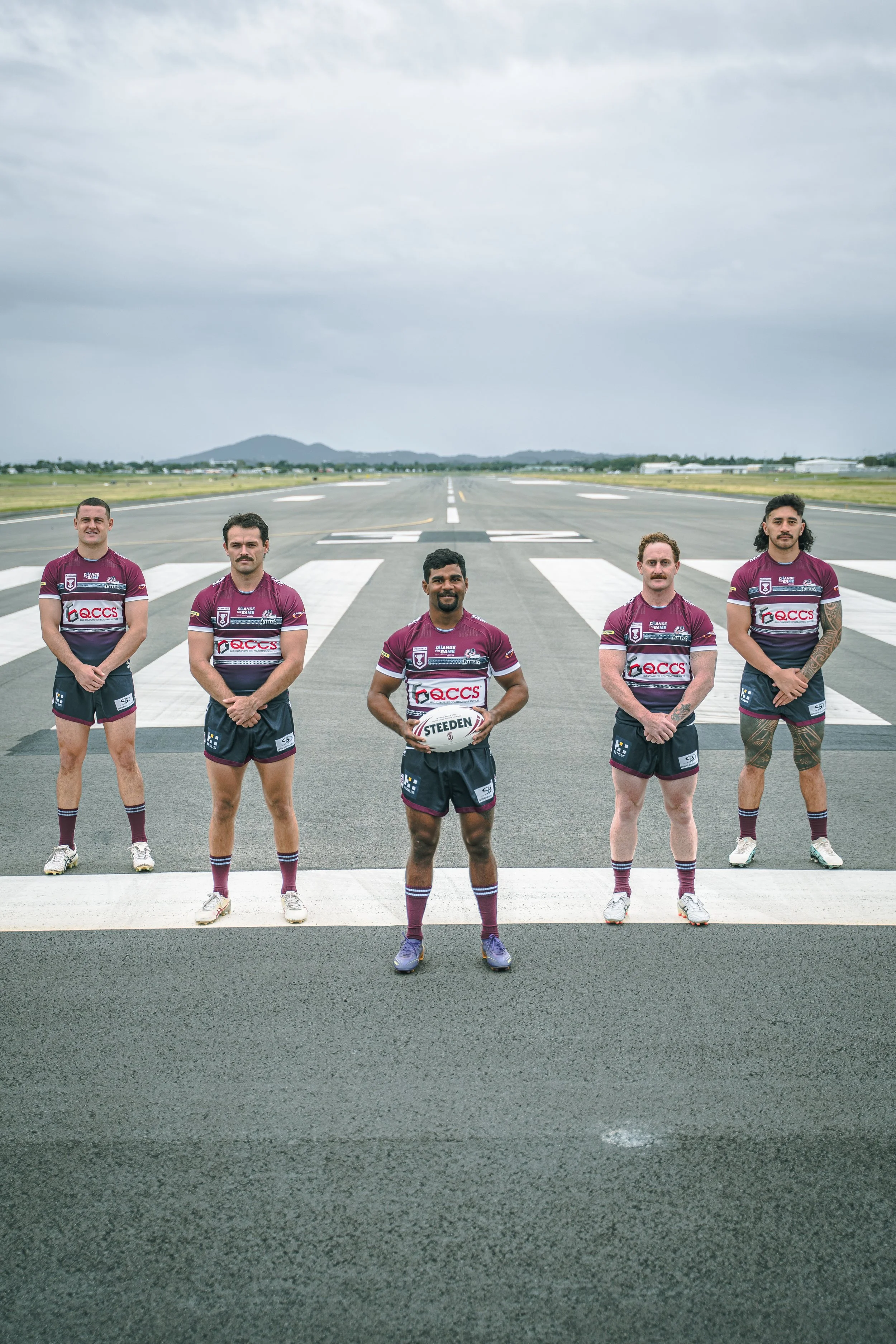 Five rugby players standing on a runway, wearing maroon jerseys and shorts, with the middle player holding a rugby ball, under a cloudy sky.