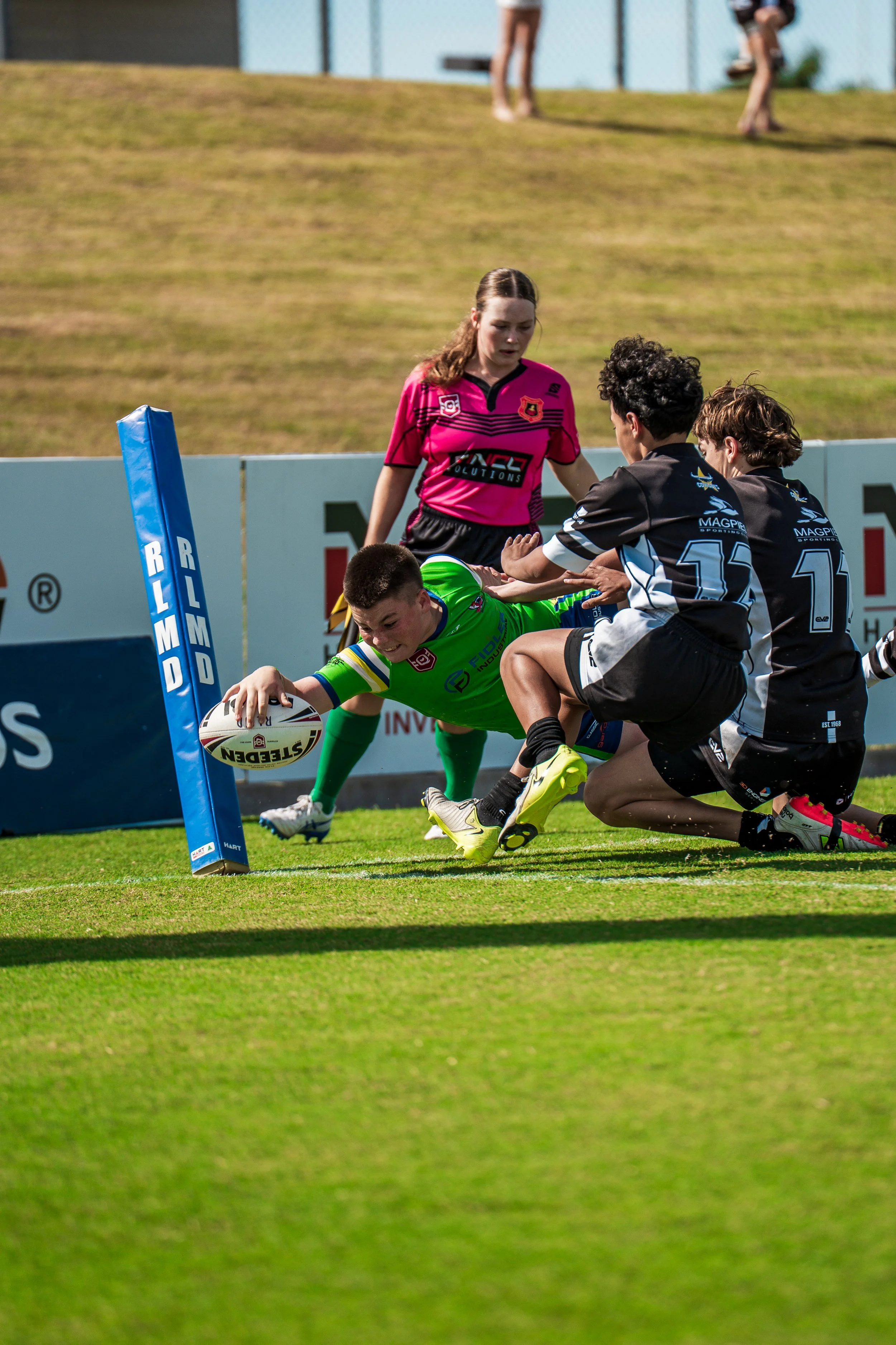 Rugby players competing for the ball near the goal post on a grassy field, with one player reaching out for the ball while others try to stop him, and a referee observing nearby.