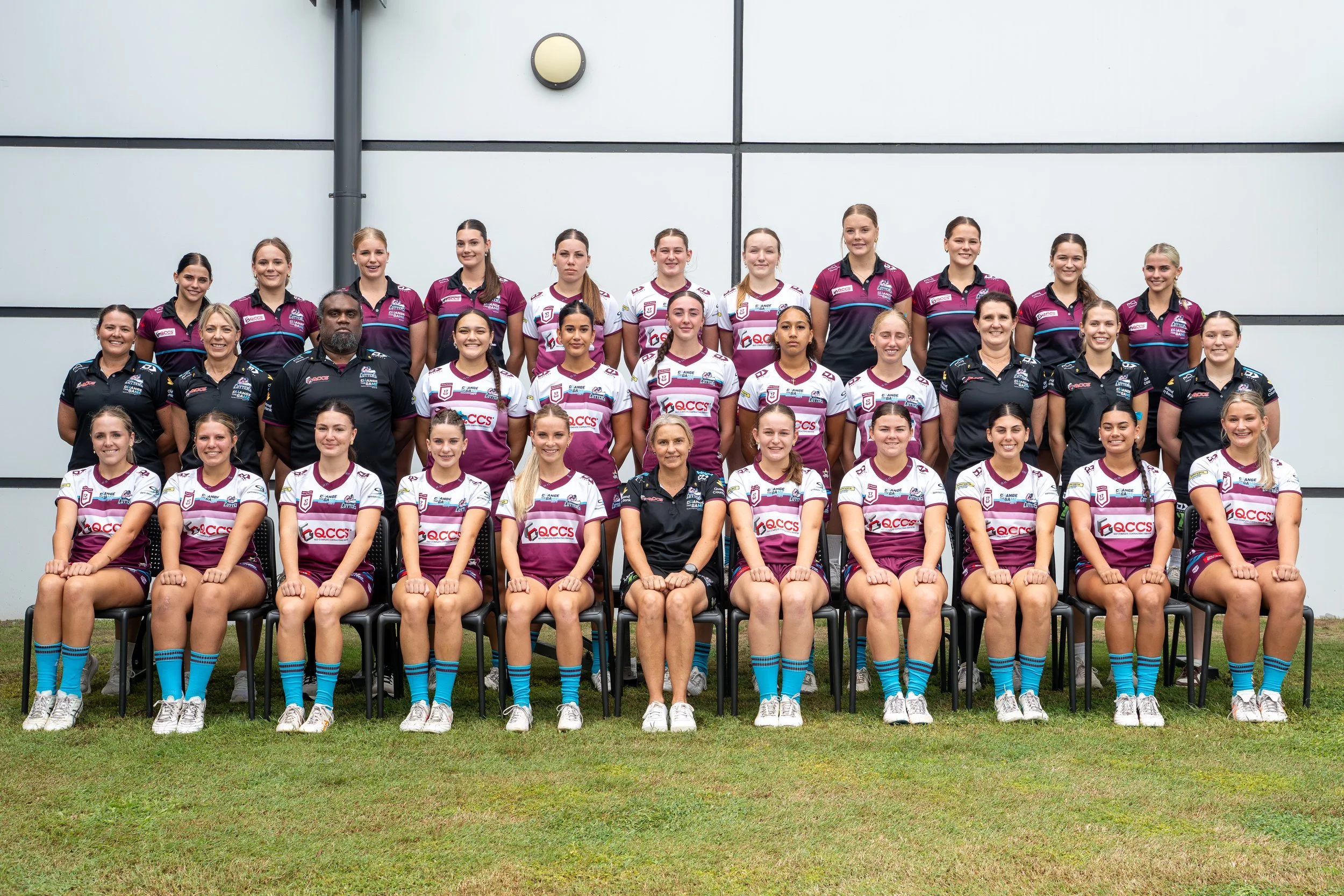 A group photo of a women's sports team with coaches, posing outdoors against a gray wall.