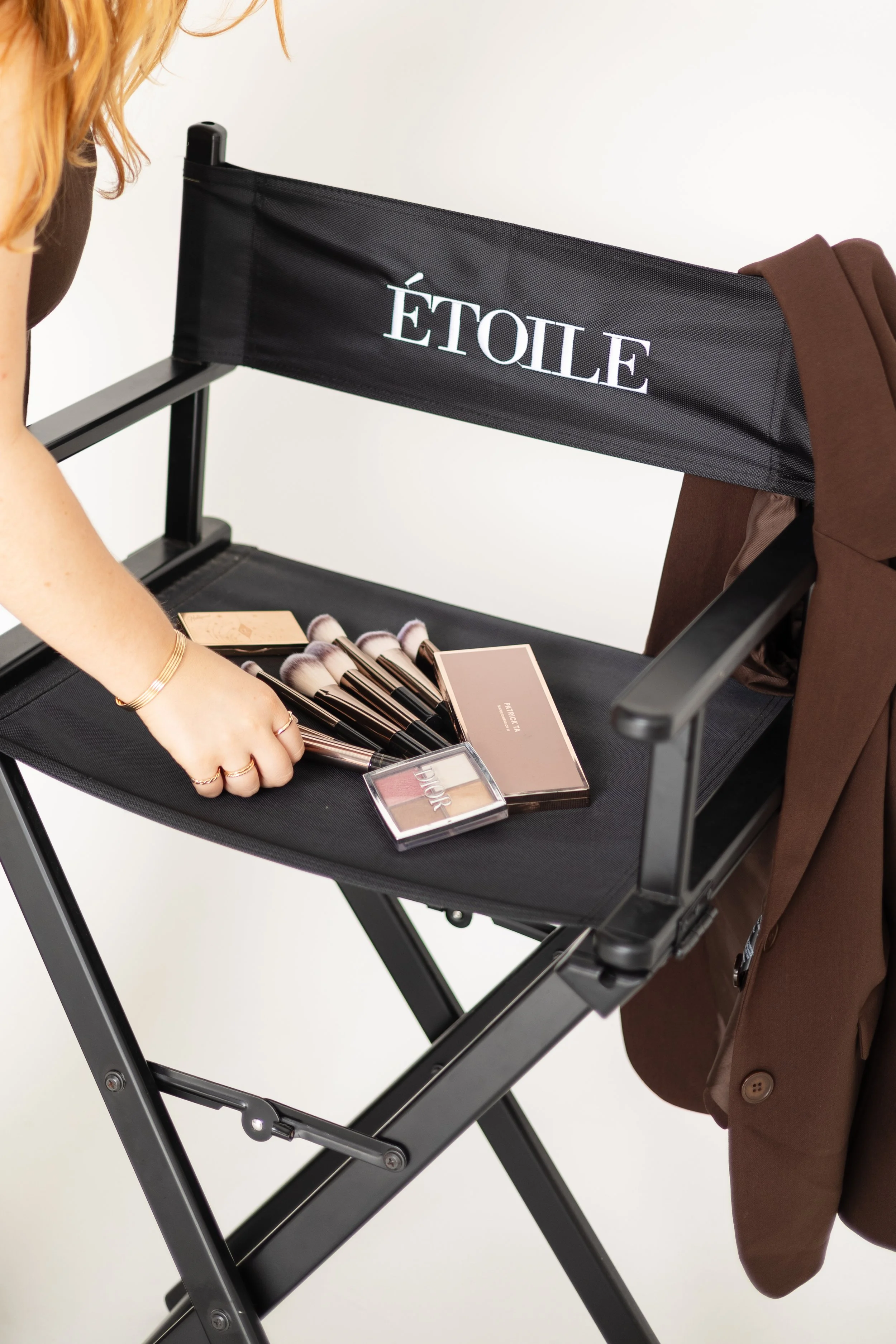A person with gold jewelry looking at makeup products arranged on a black foldable chair with a brown blazer draped over the back, and the chair has a logo that reads 'ÉTOILE'.