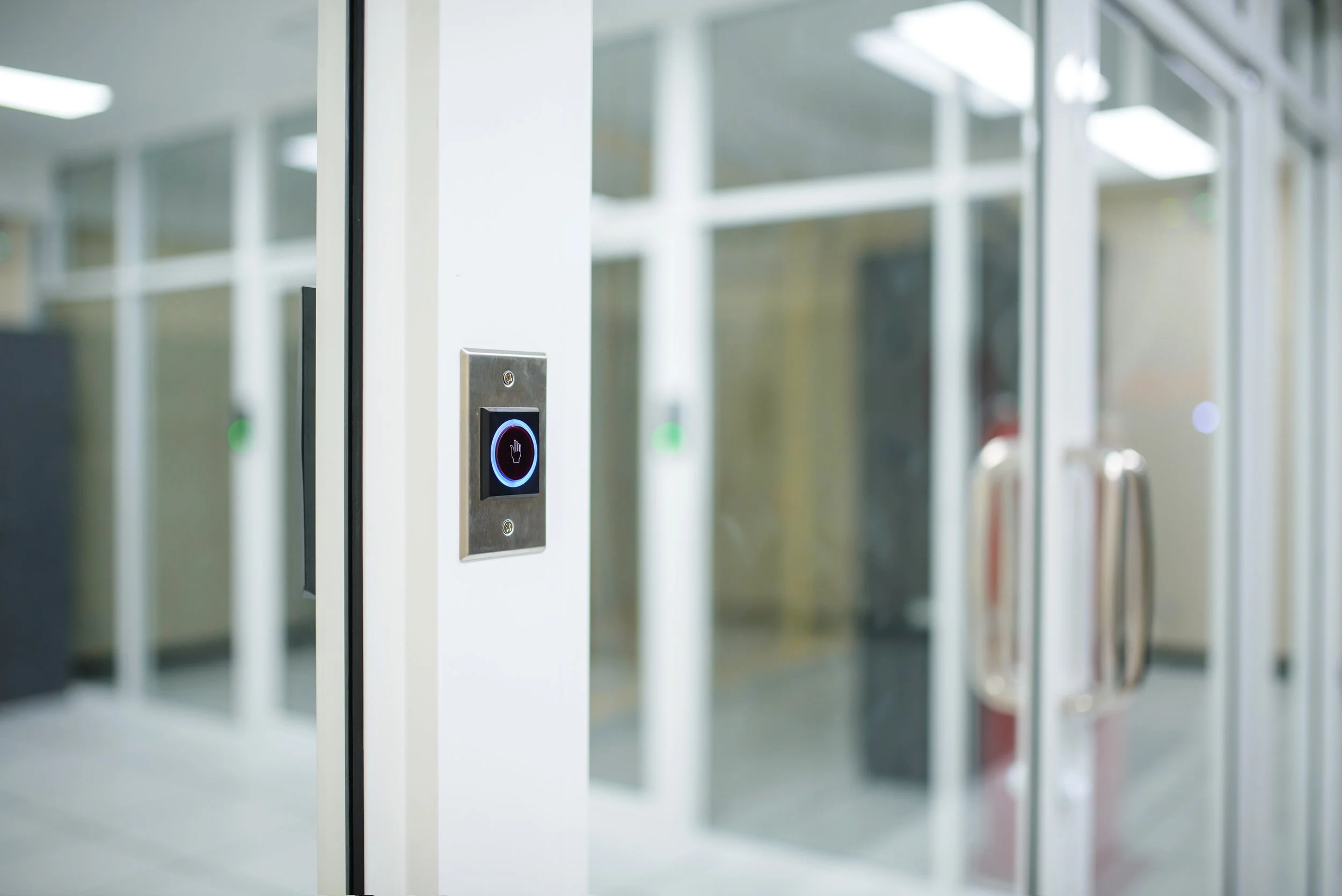 Close-up of a modern elevator call button with a red hand symbol for stop or hold, and an illuminated blue circle, in a building with glass doors and reflections.