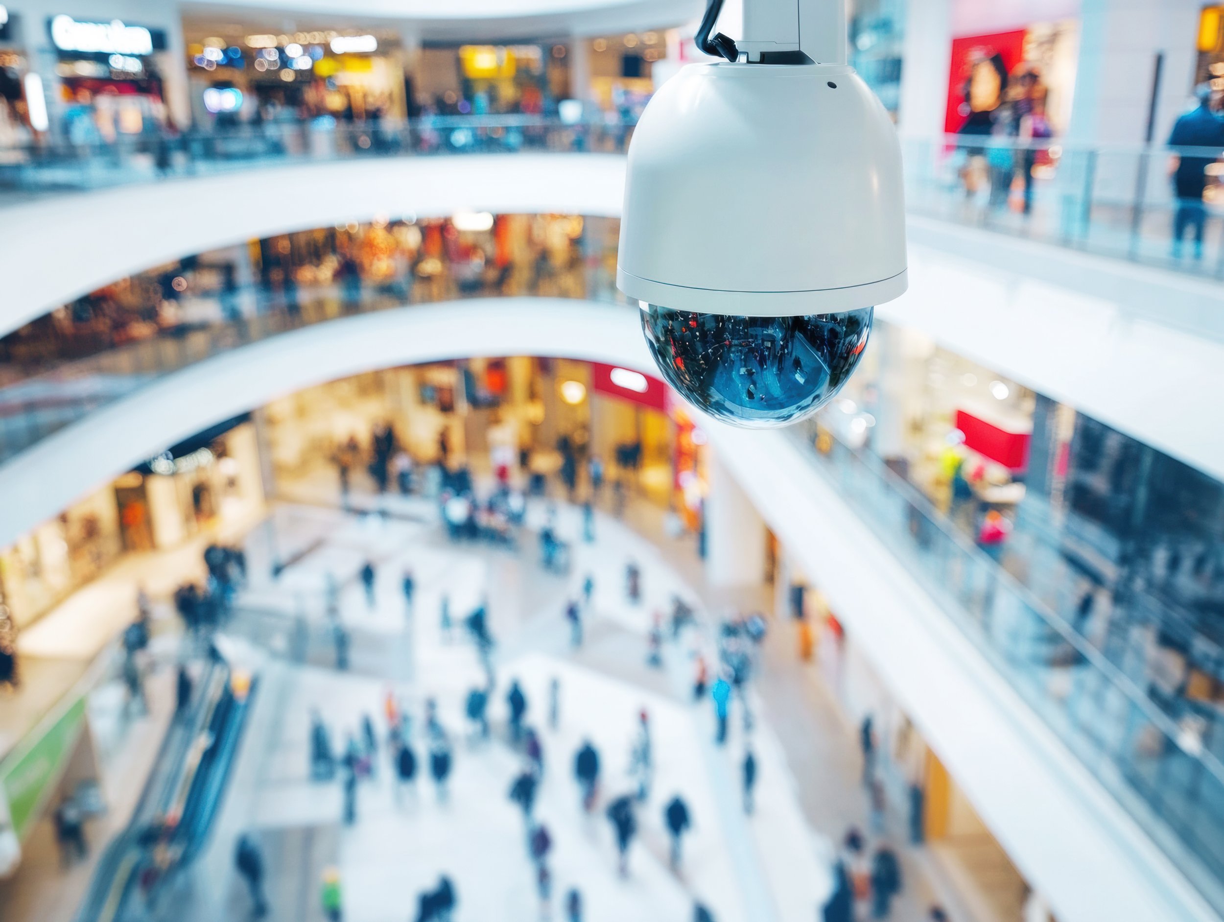 Security camera hanging from the ceiling inside a multi-level shopping mall with blurred shoppers and store signs in the background.