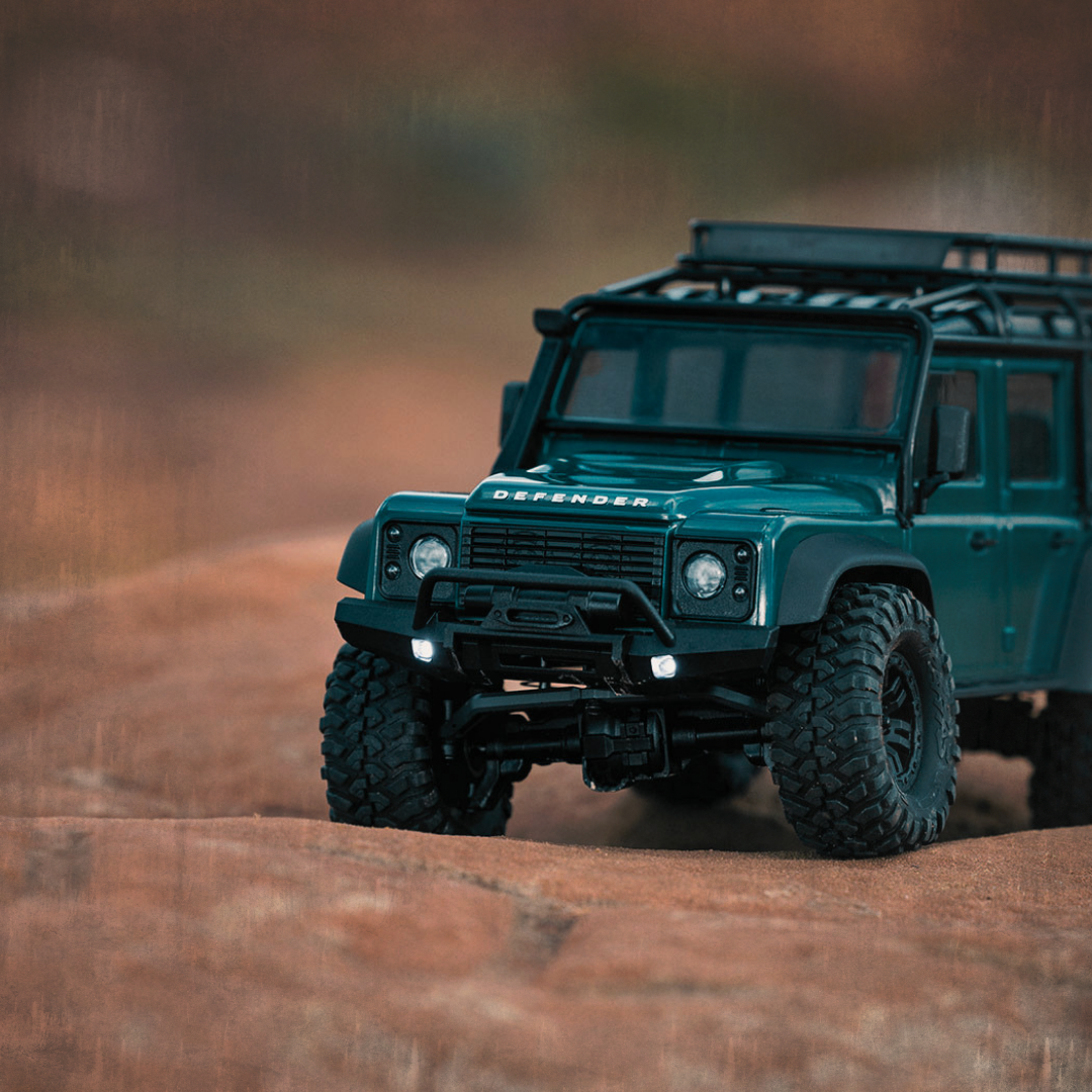A detailed close-up of a scale model of a teal Land Rover Defender on a rocky surface, with a blurred natural background.