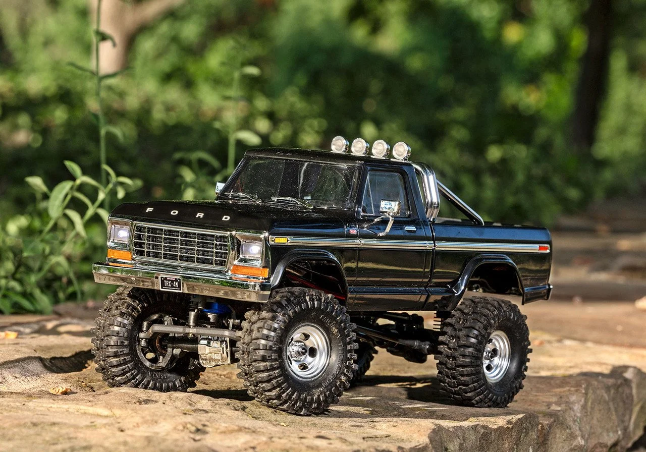 A black toy monster truck with large tires and shiny chrome details, positioned outdoors on a dirt surface with green foliage in the background.