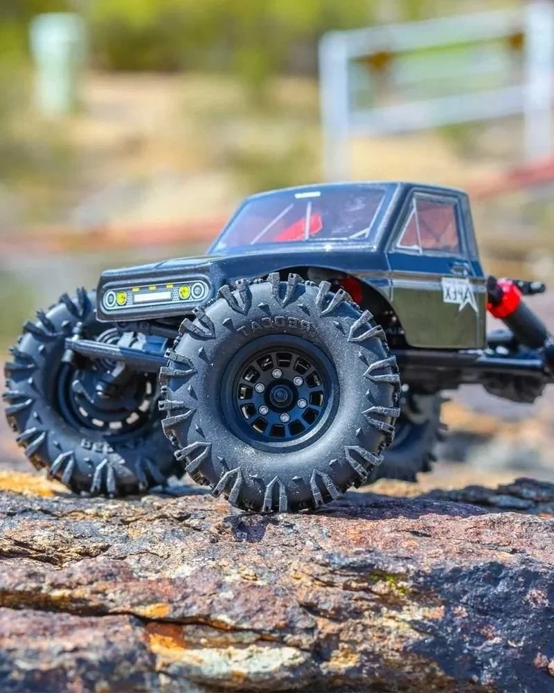 Close-up of a RedCat Ascent-18 APEX off-road truck with large rugged tires on a rocky surface, background of blurred greenery and structures.