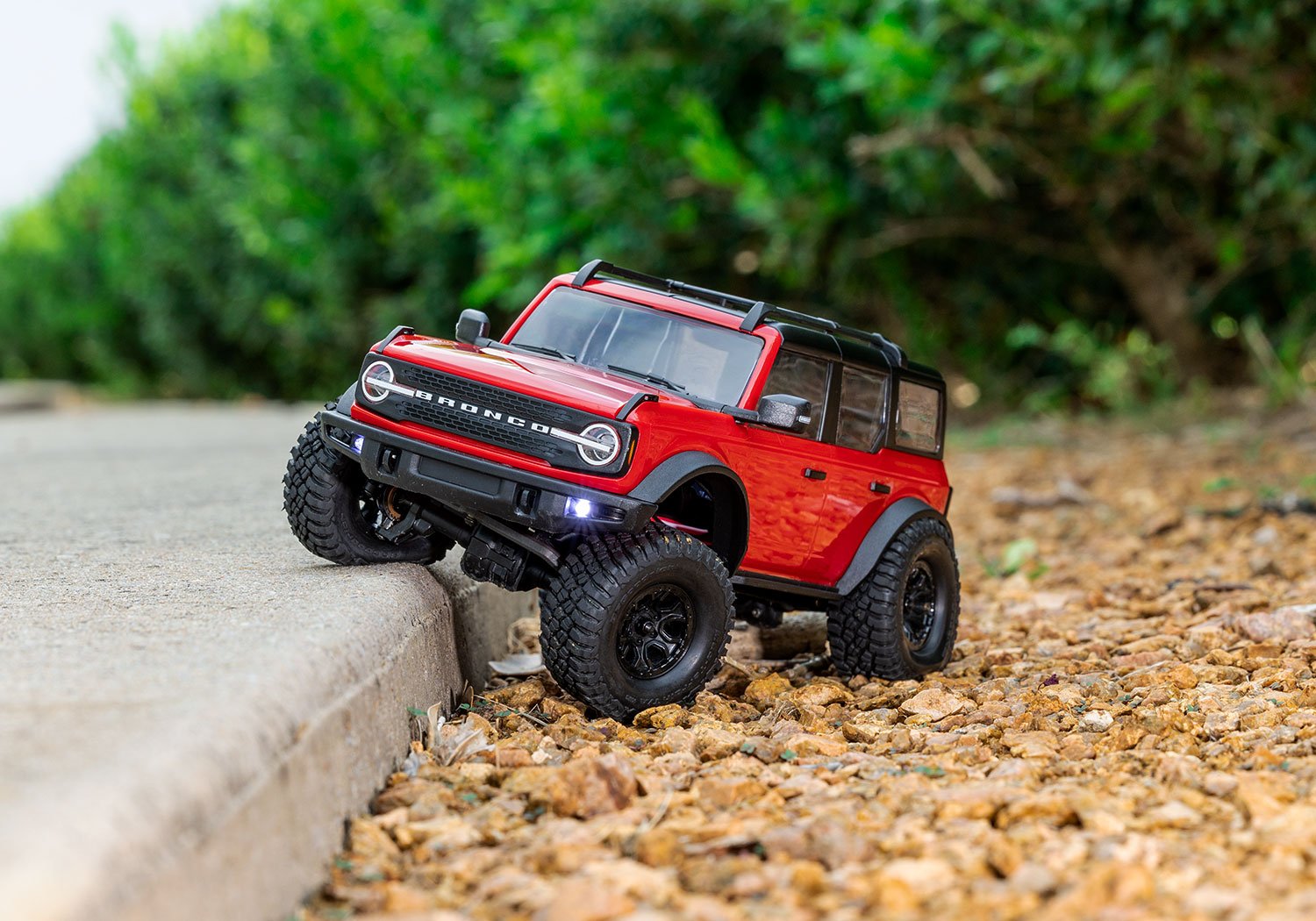 A red remote-controlled toy car modeled after a Ford Bronco on a gravel surface near a curb with green bushes in the background.