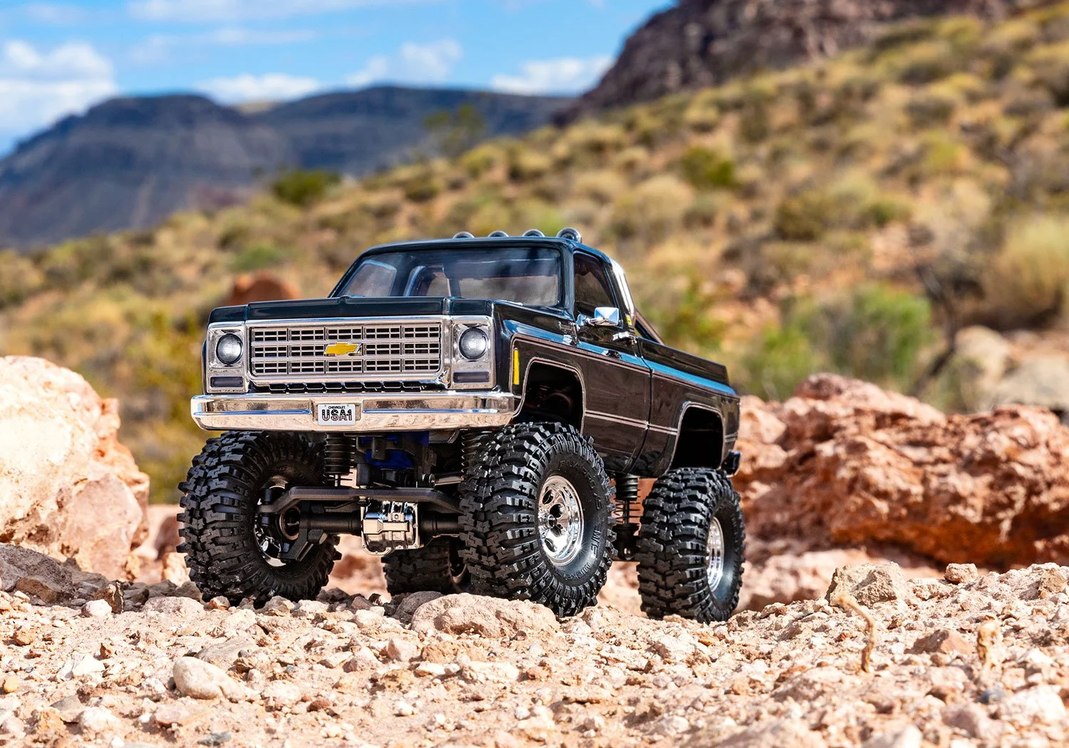 A detailed remote-controlled toy pickup truck with large off-road tires on rocky terrain in a desert landscape with mountains and blue sky in the background.