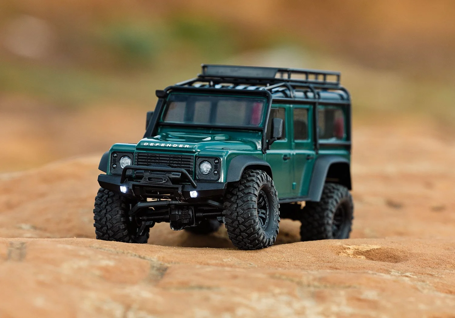 A detailed model of a green Land Rover Defender with black tires and accessories, positioned on a sandy surface.