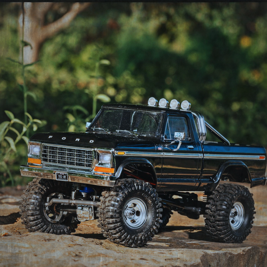 A black toy pickup truck with rugged tires and off-road suspension, placed on a rocky surface outdoors, with green foliage in the background.